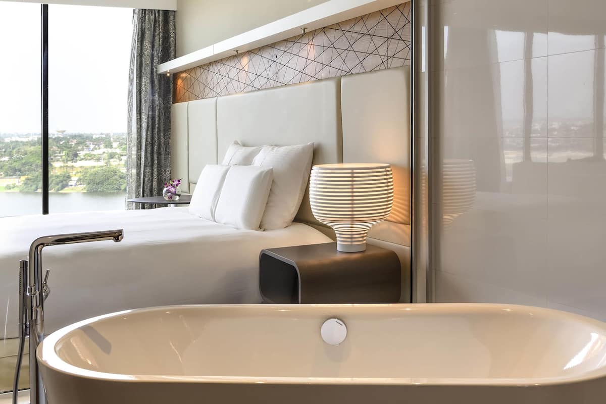A modern bathroom with a freestanding bathtub, a bedside table with a lamp, and a view of a cityscape through large windows.