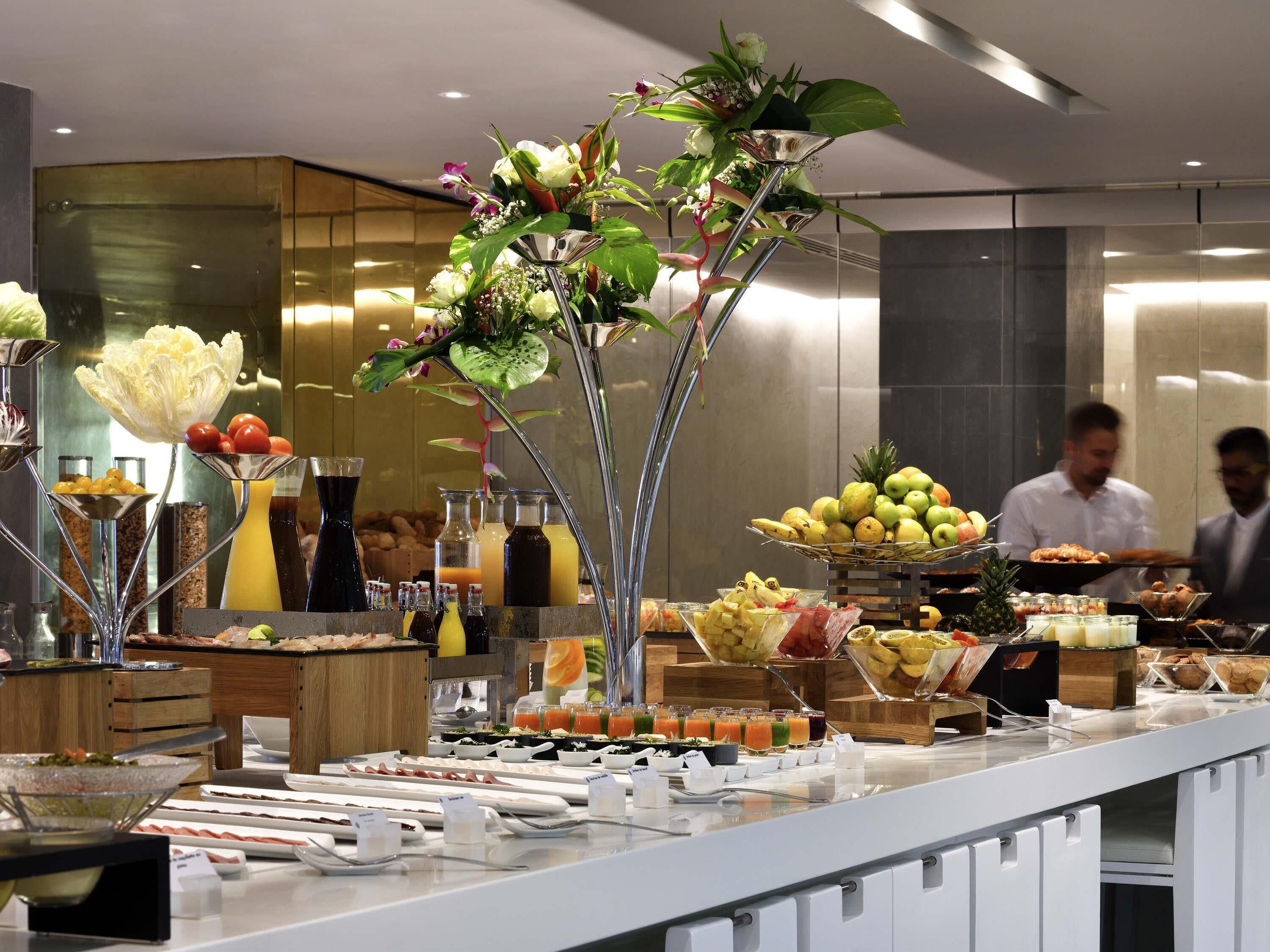 A buffet with various food items and drinks, including fruits, vegetables, and a floral centerpiece.