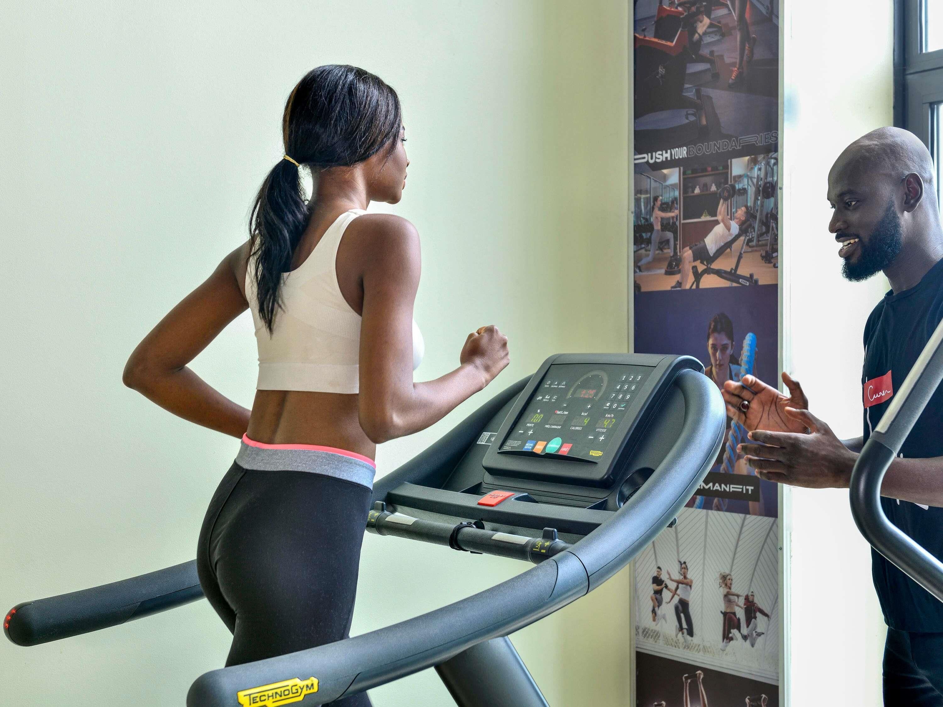 A woman running on a treadmill in a gym with a man standing beside her.