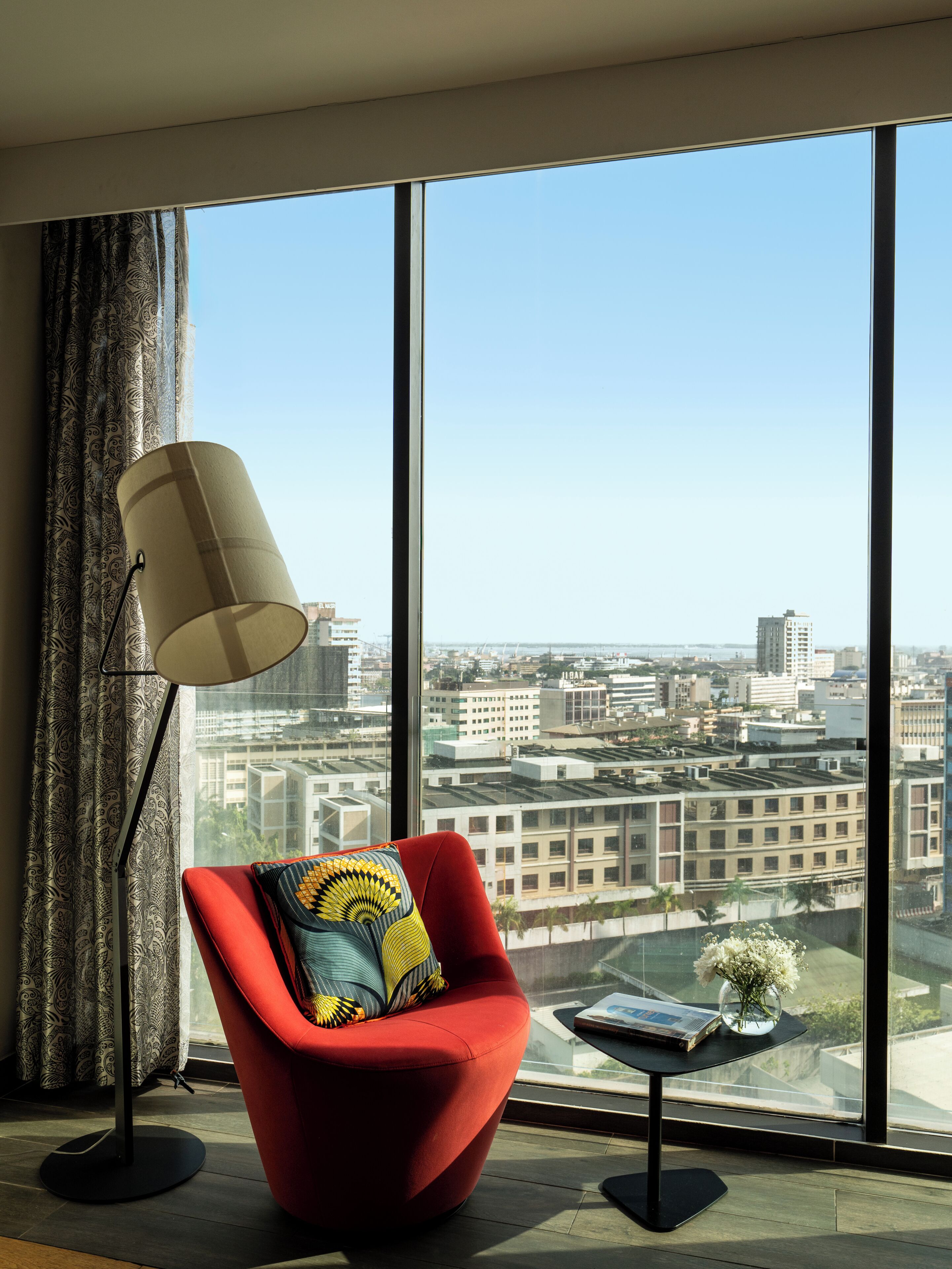 A modern room with a large window, a red chair, a floor lamp, and a small table with a vase.