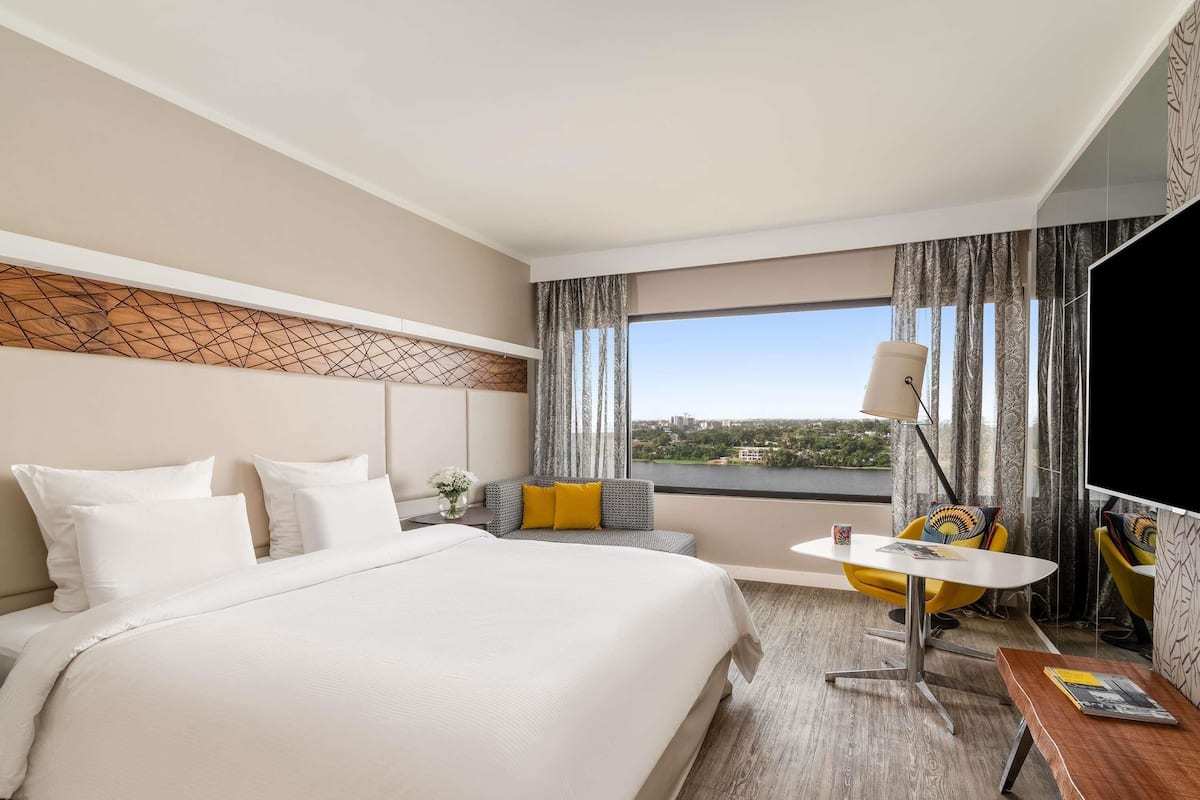 A hotel room with a large bed, a small sofa, a table, and a view of a cityscape through the window.