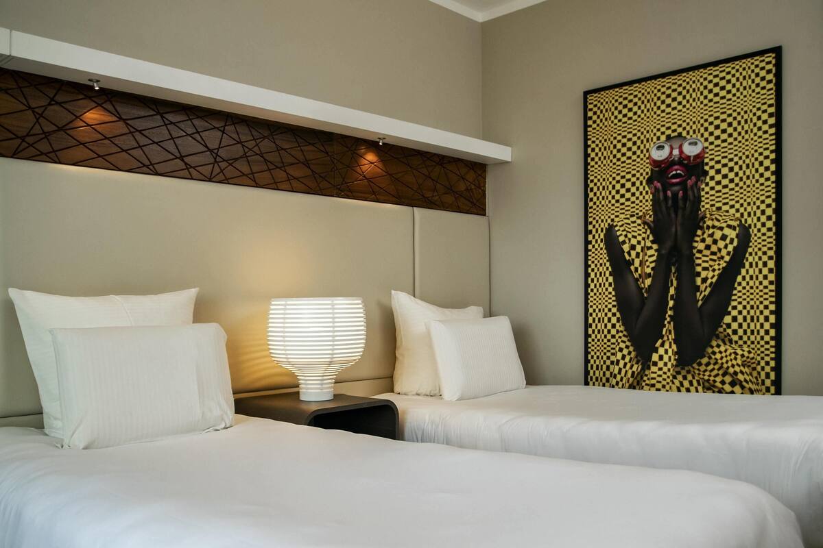 A hotel room with two beds, a nightstand, a lamp, and a wall-mounted artwork.