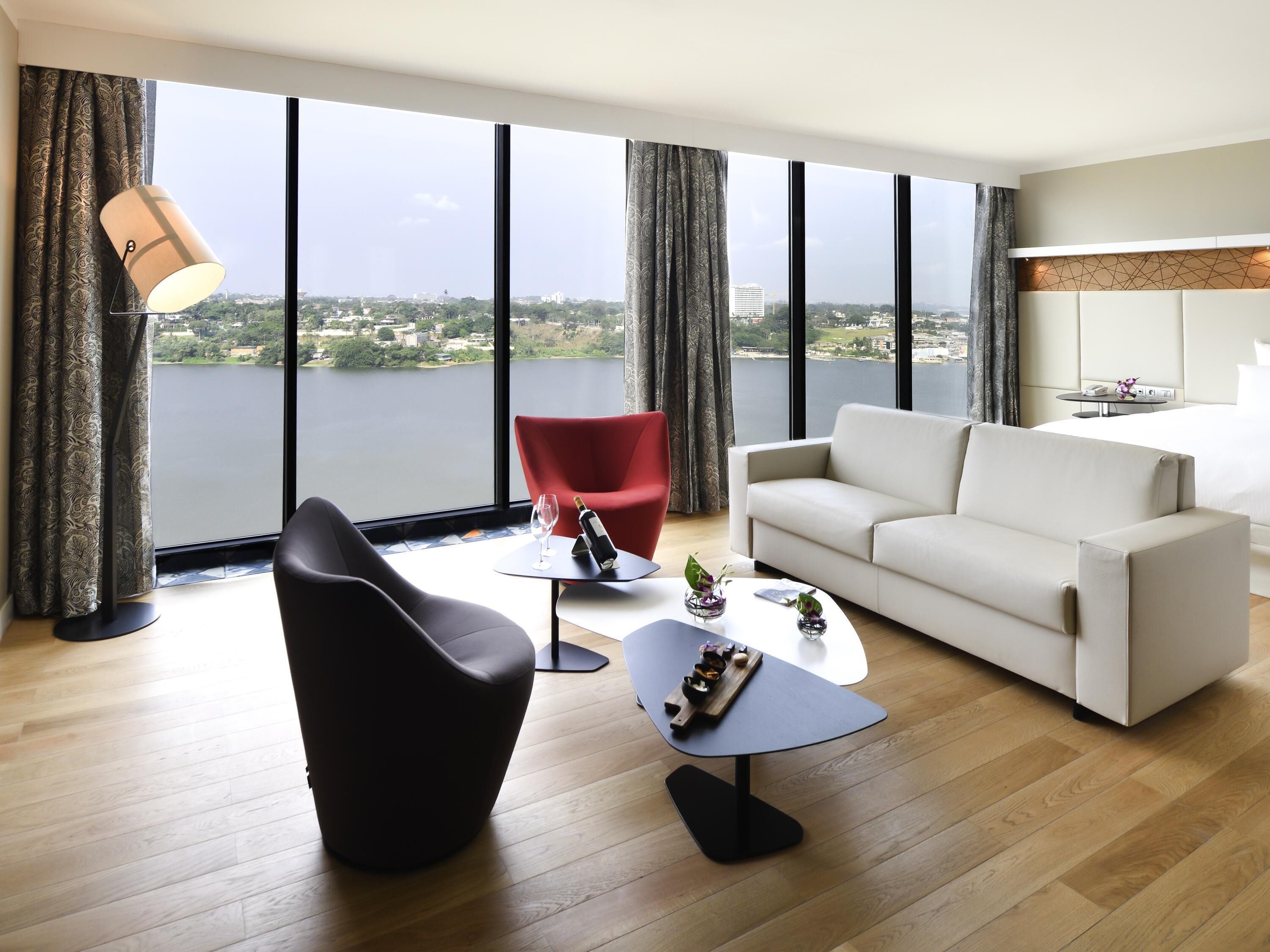 A modern living room with a large window, a red chair, a white sofa, and a small table with a bottle and glass.