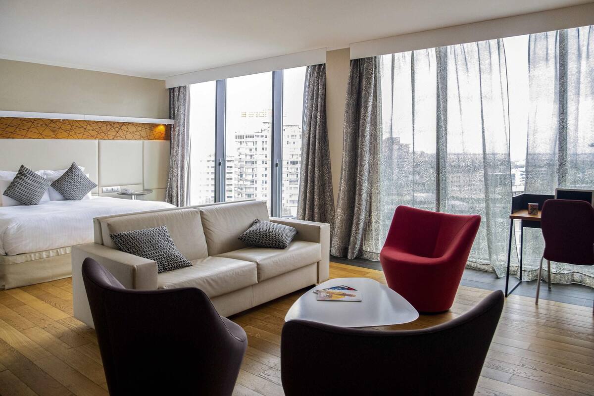 A modern hotel room with a large bed, a sofa, two armchairs, a small table, and a view of the city through large windows.