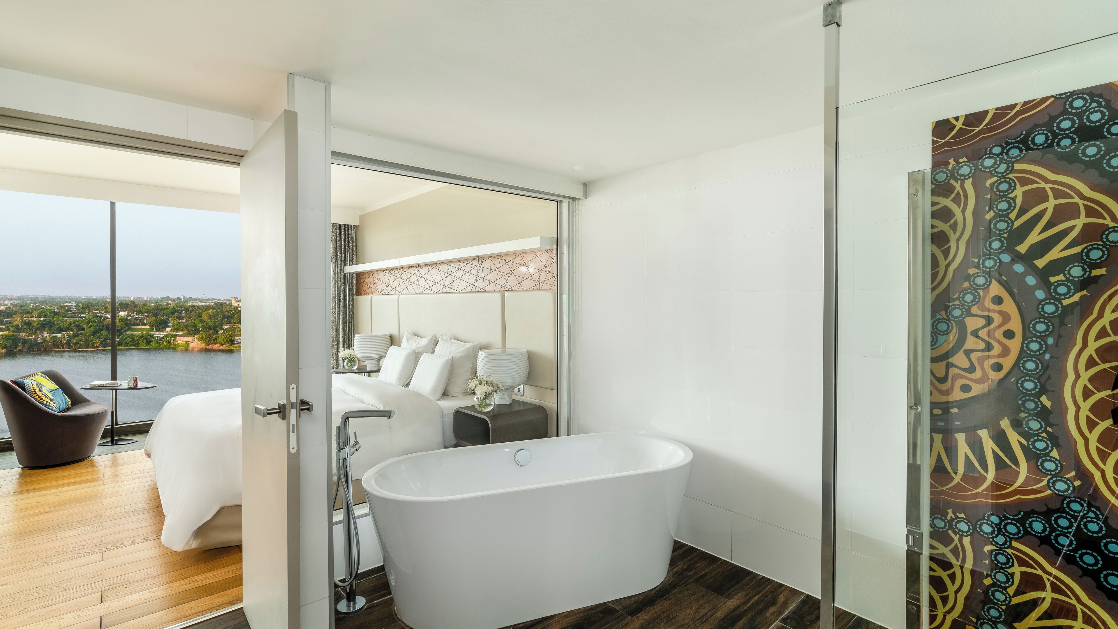 A modern bathroom with a freestanding bathtub, a glass shower enclosure, and a view of a bedroom with a large bed and a cityscape.