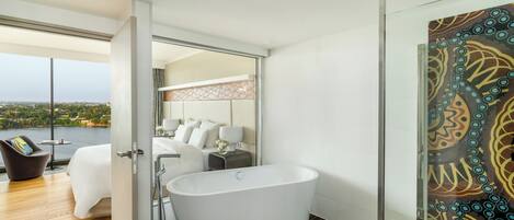 A modern bathroom with a freestanding bathtub, a glass shower enclosure, and a view of a bedroom with a large bed and a cityscape.