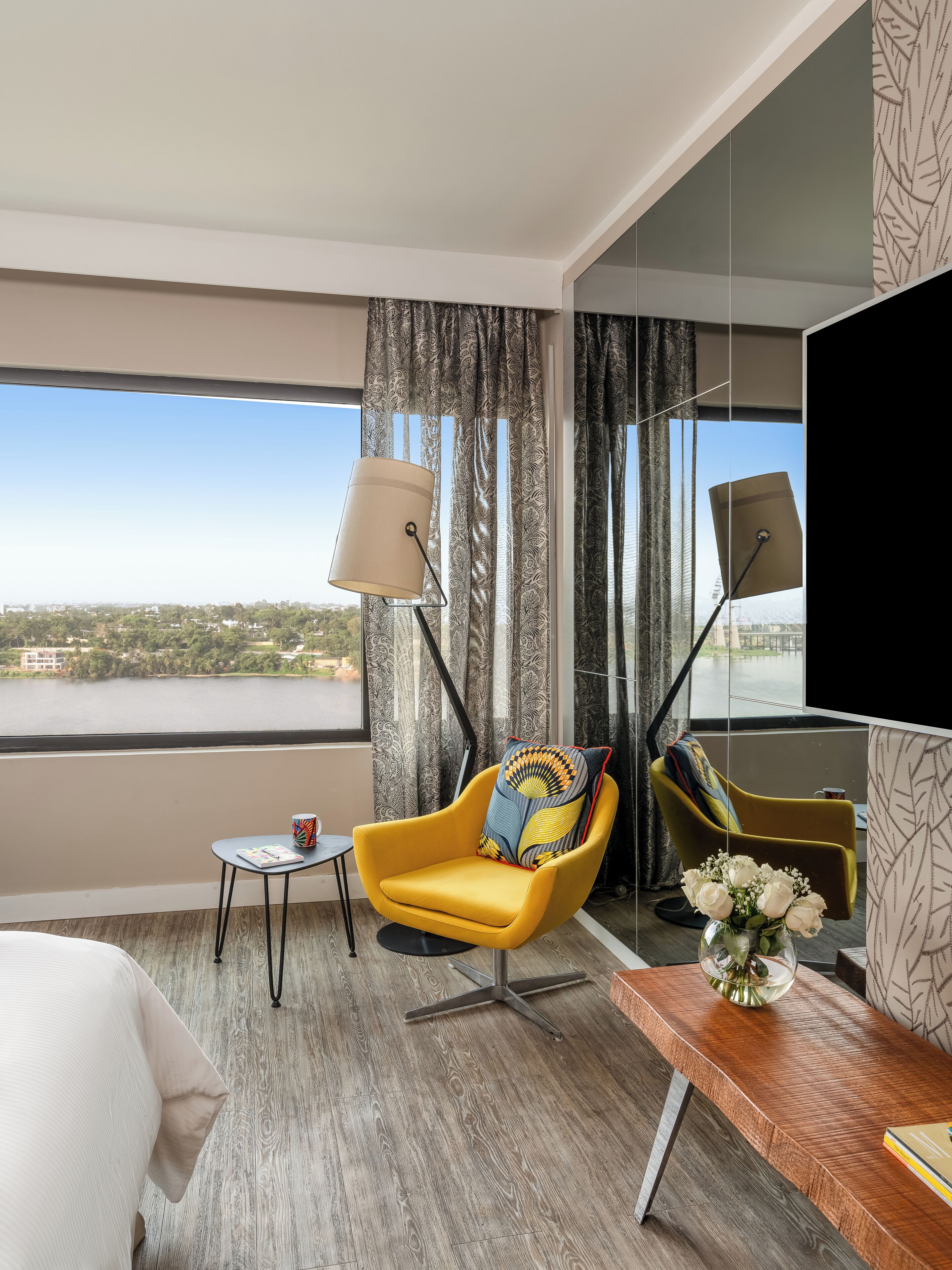 A hotel room with a yellow armchair, a small table with a cup, a large window with a view of a body of water, and a flat-screen TV mounted on the wall.