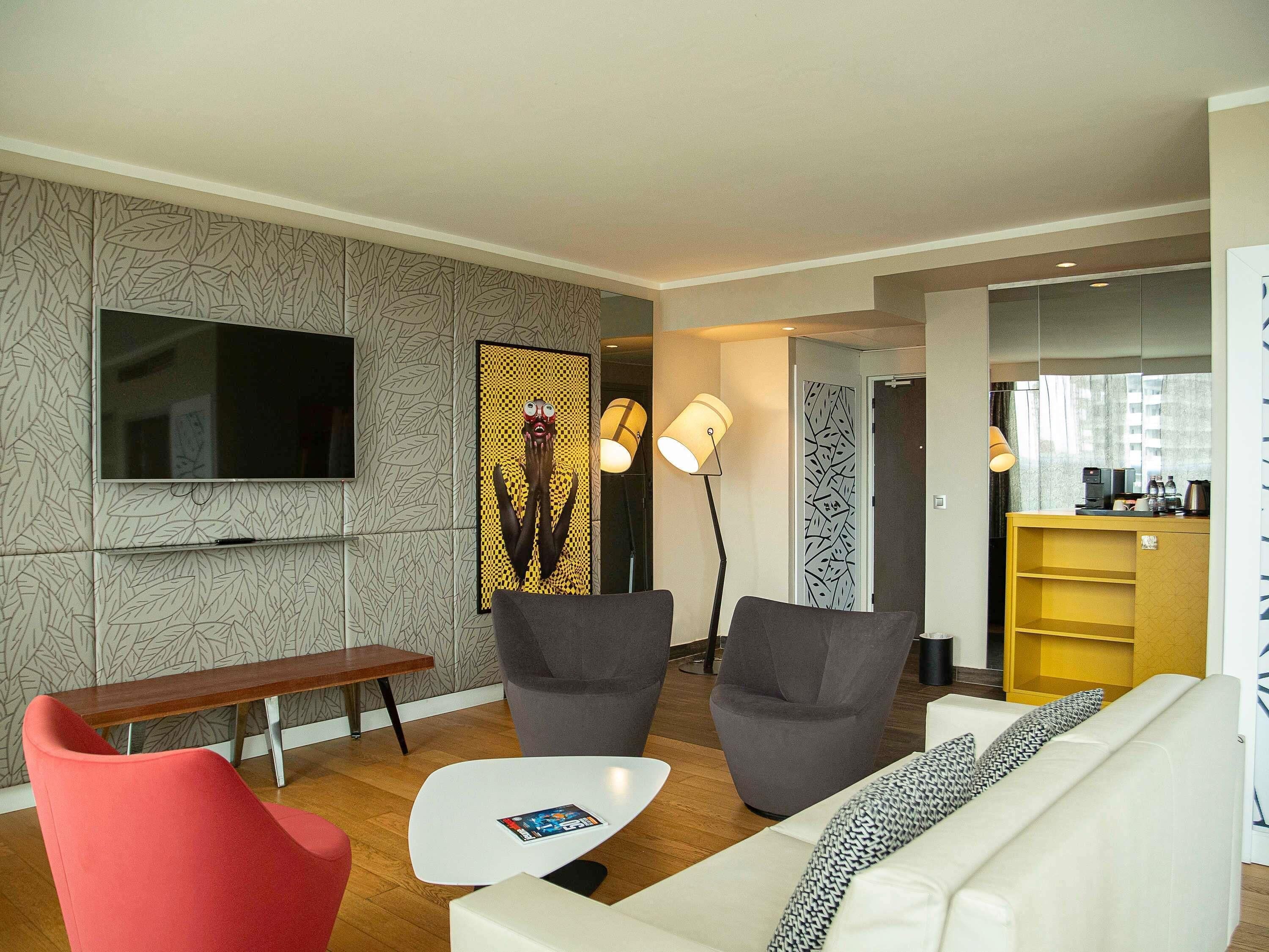 Un salon moderne avec un téléviseur à écran plat, deux fauteuils gris, un fauteuil rouge, un canapé blanc, une petite table blanche et un coin café.