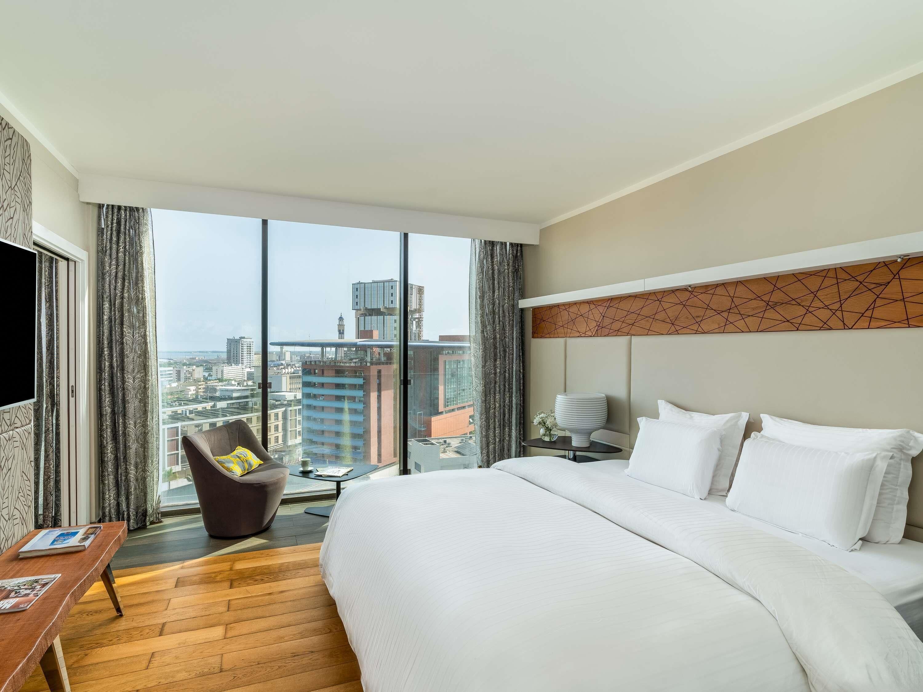A hotel room with a large bed, a desk, a chair, and a view of the cityscape.