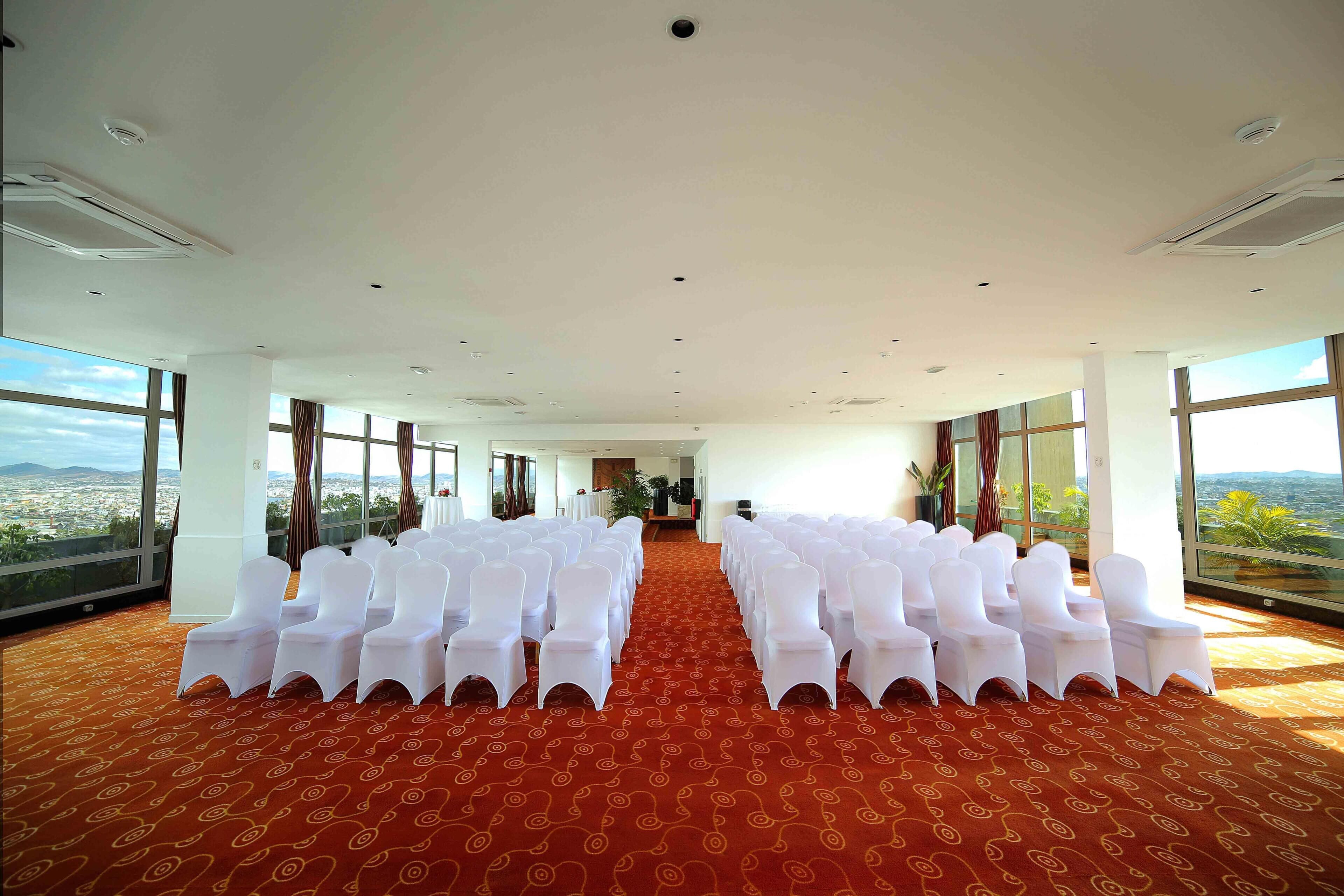 Un salón de conferencias con filas de sillas blancas, ventanas amplias y una alfombra roja.
