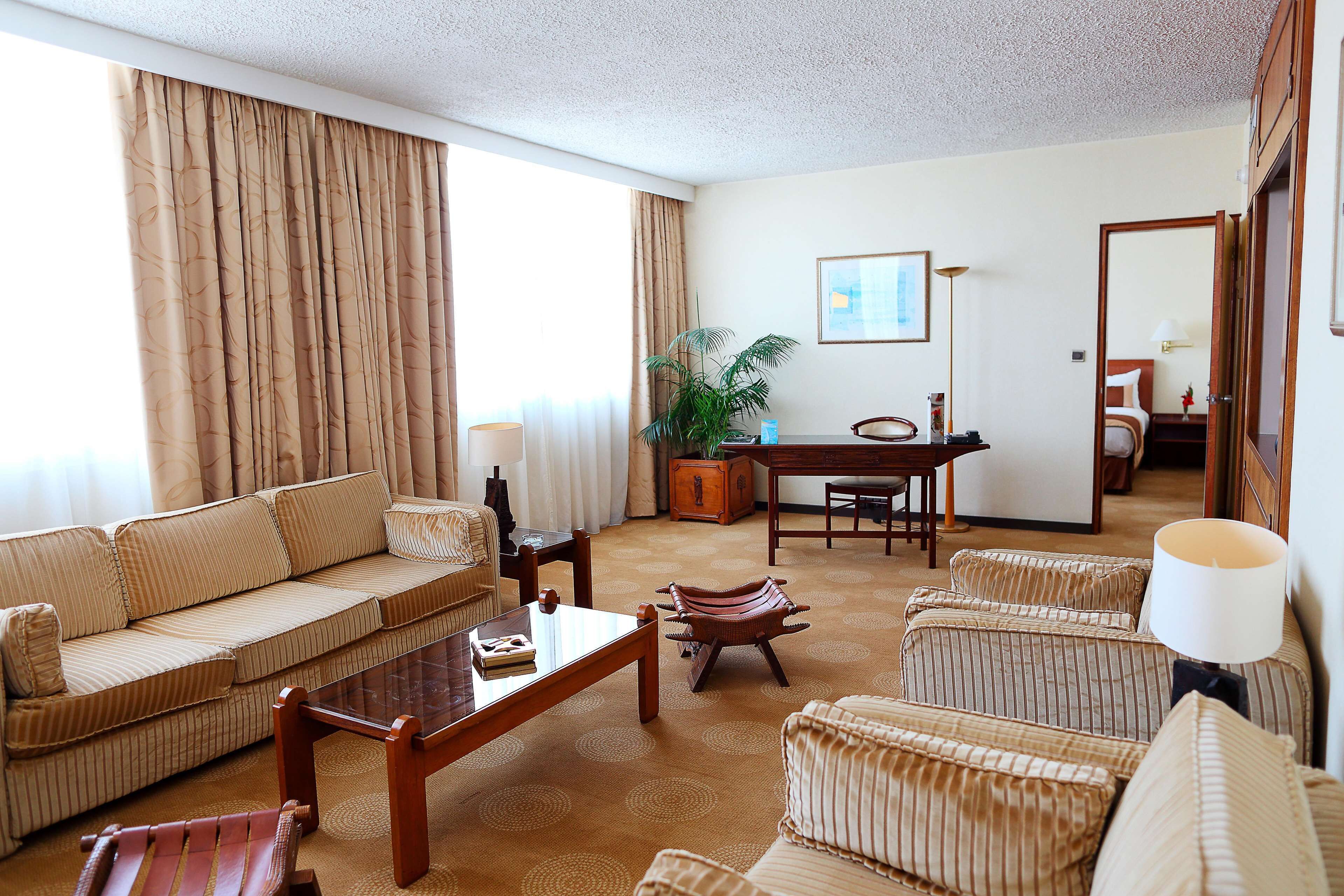 A hotel room with a sofa, armchairs, a dining table, a lamp, and a potted plant.