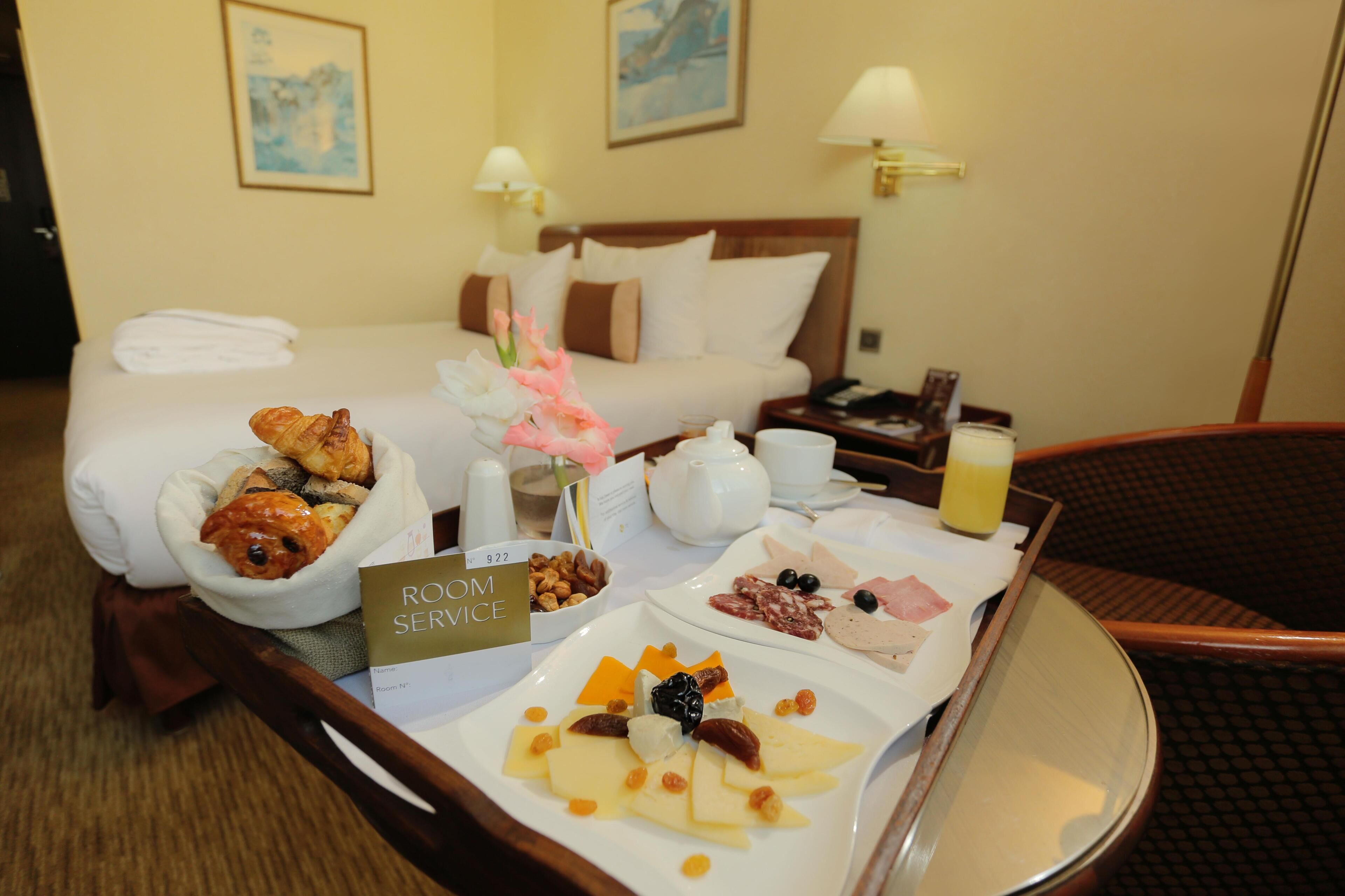 Una camera d'albergo con un letto, un comodino con una lampada, un vassoio con prodotti per la colazione e un cartello con scritto 'ROOM SERVICE'.