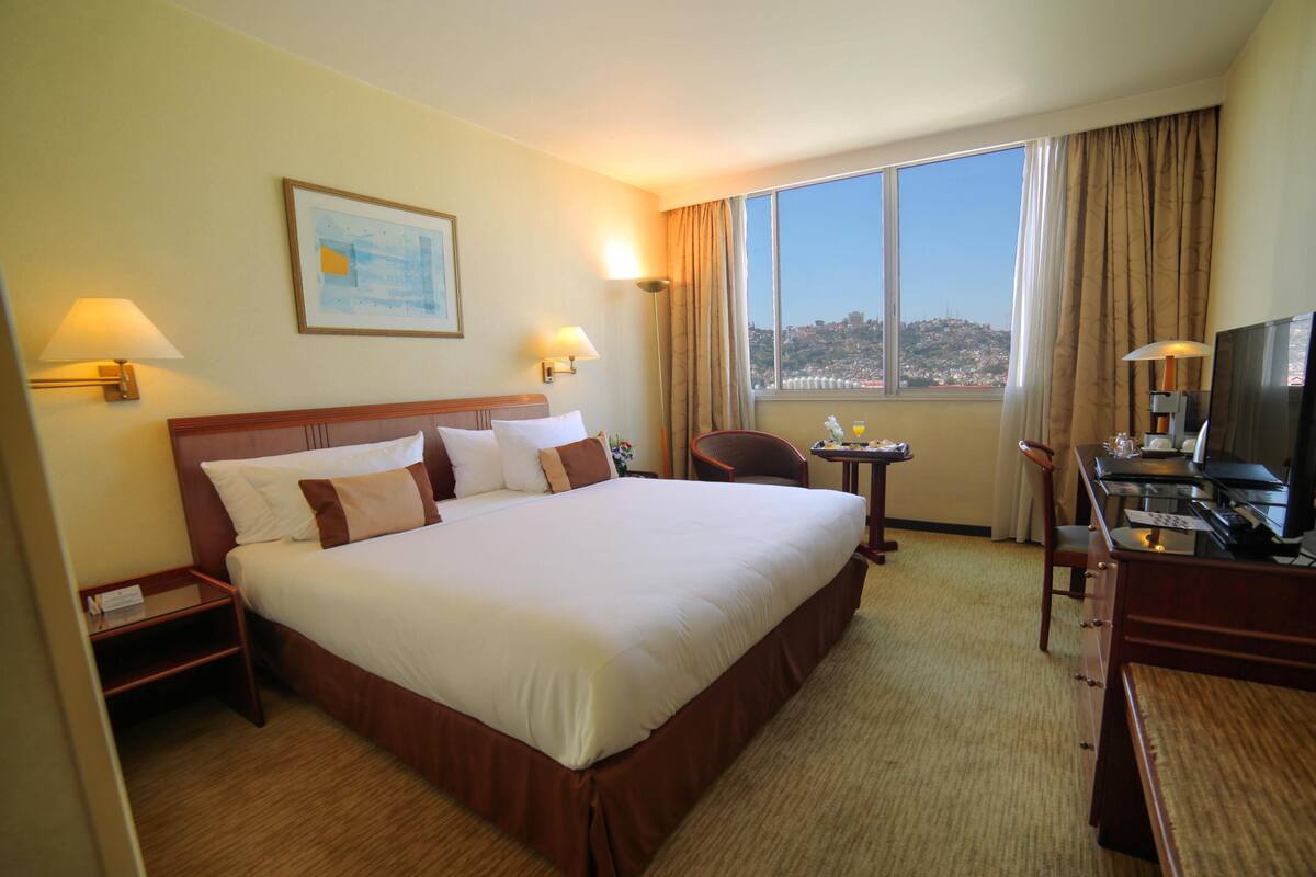 A hotel room with a large bed, two bedside lamps, a desk with a TV, and a view of the city.