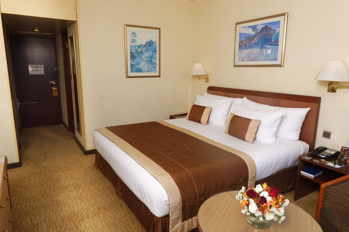 A hotel room with a large bed, two wall-mounted lamps, a bedside table with a phone and a book, and a small round table with a vase of flowers.