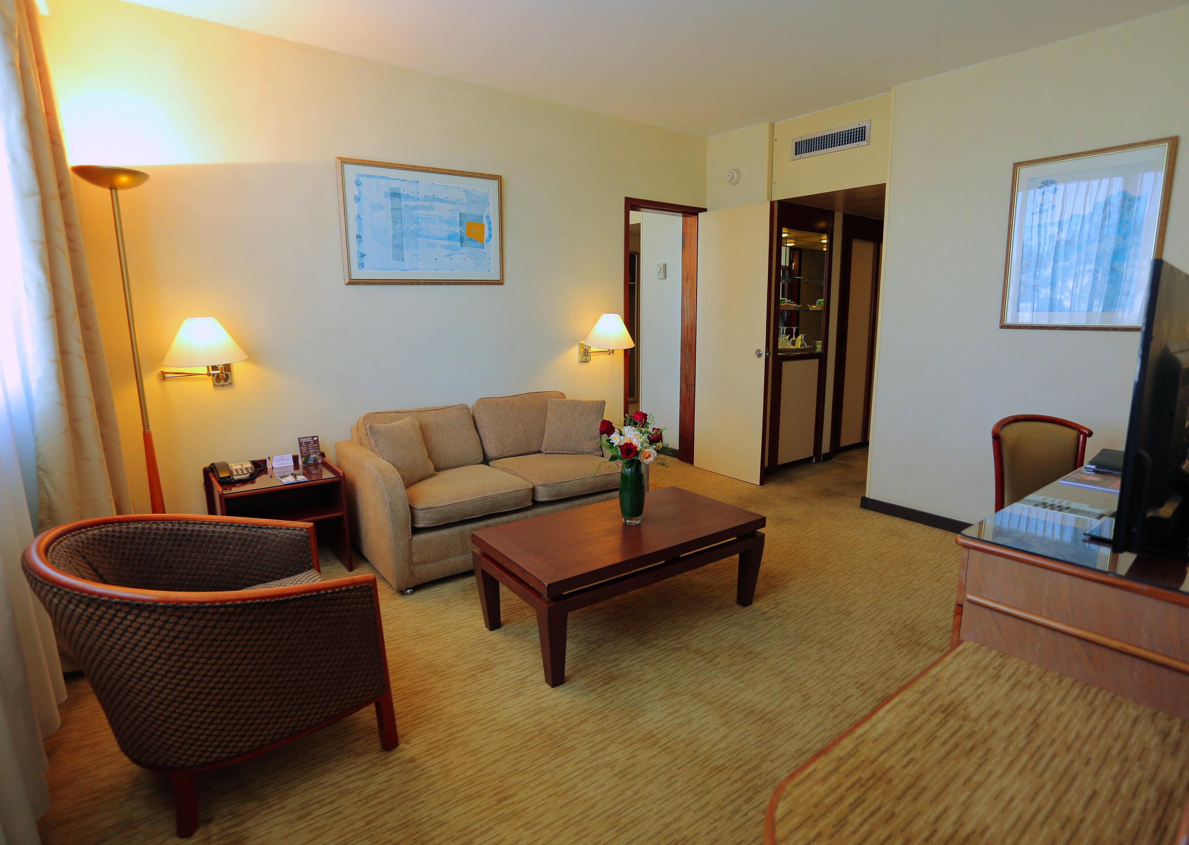 A hotel room with a sofa, armchair, coffee table, TV, lamp, and a painting on the wall.