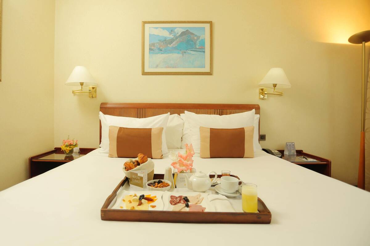 A hotel room with a bed, a tray with breakfast items, a painting on the wall, and two bedside lamps.