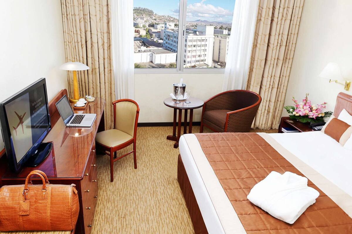 A hotel room with a bed, a desk with a laptop, a chair, a small table with a drink, a handbag, and a window with a city view.