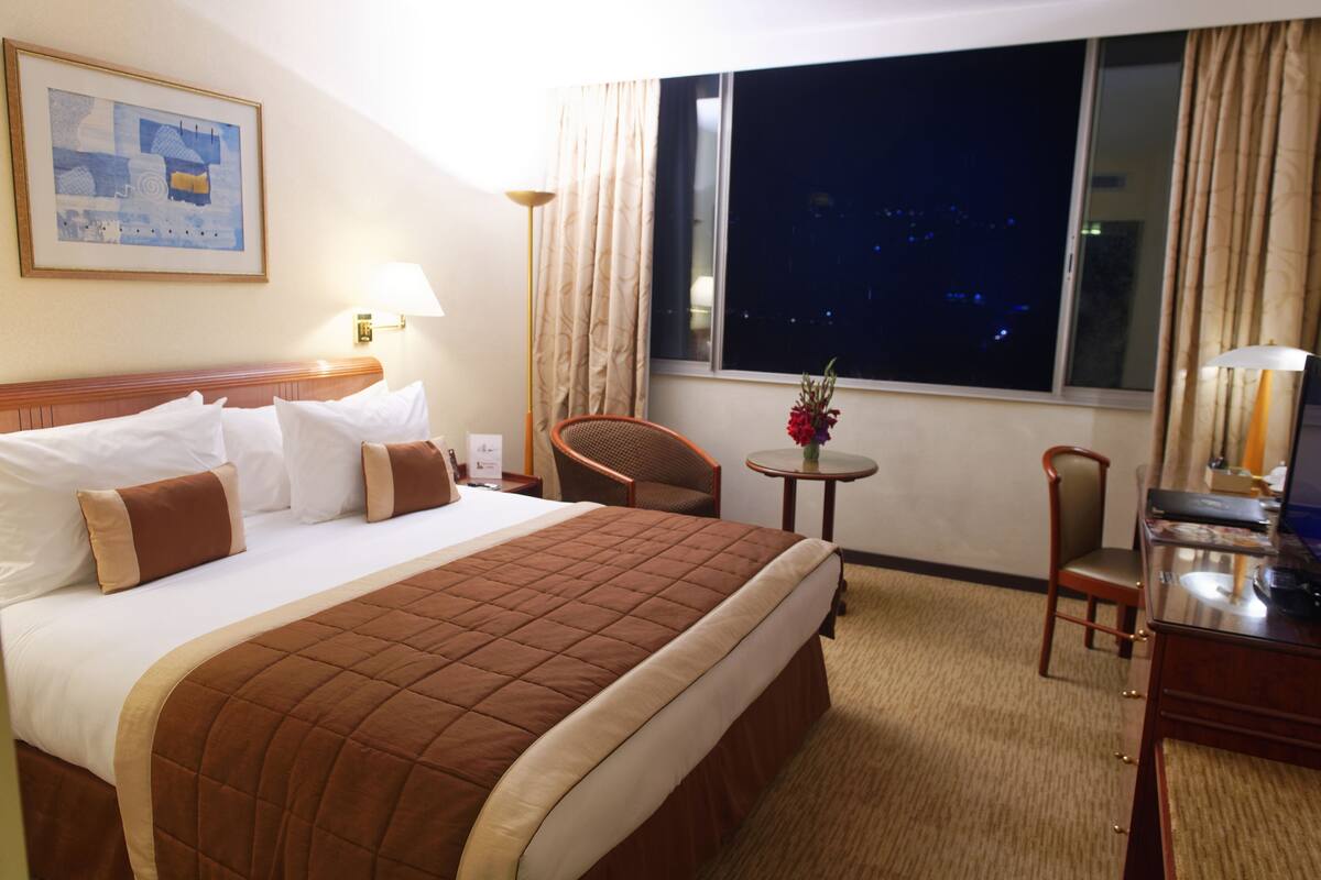 A hotel room with a bed, a chair, a small table with a vase, and a desk with a lamp.