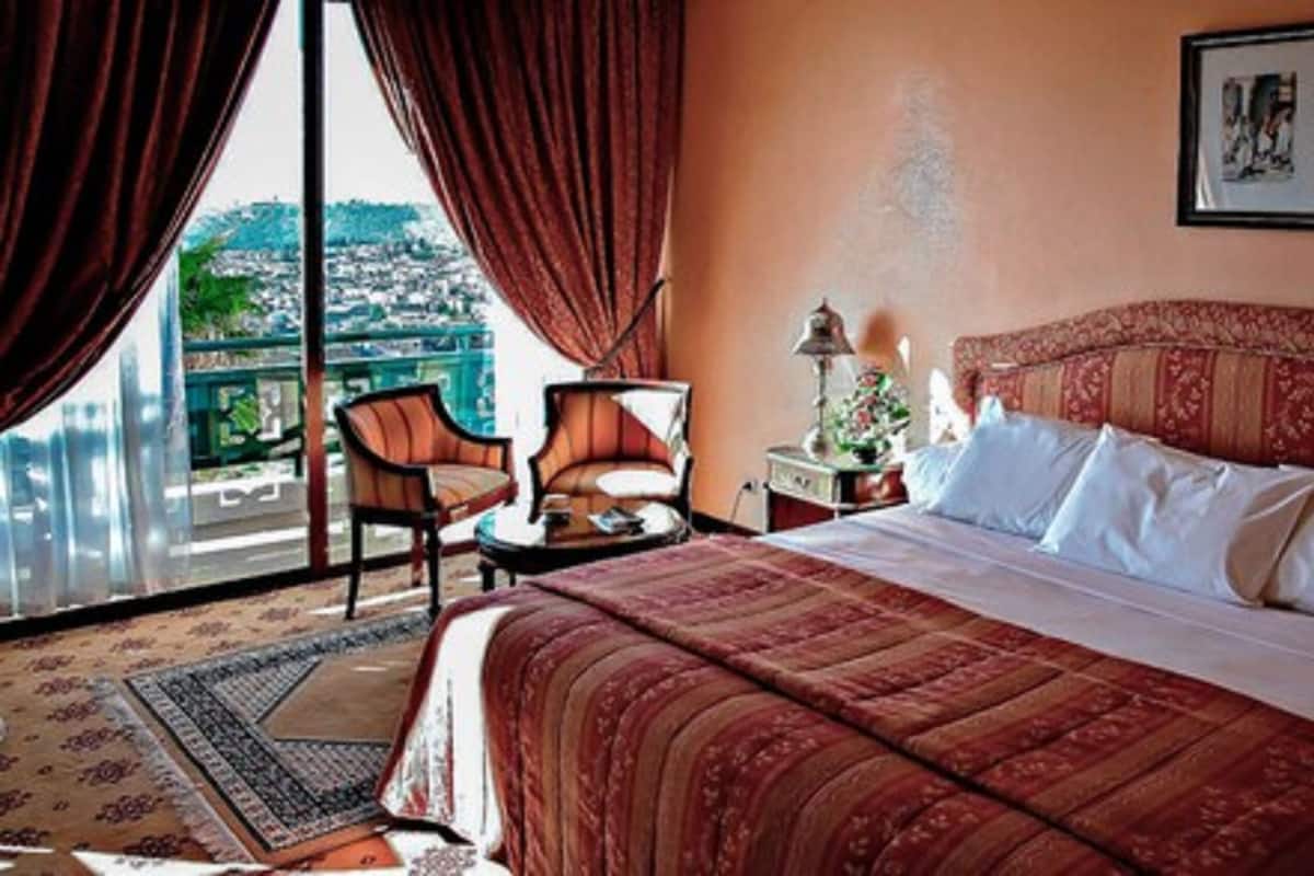 A hotel room with a large bed, two chairs, a nightstand, and a view of a cityscape through large windows.
