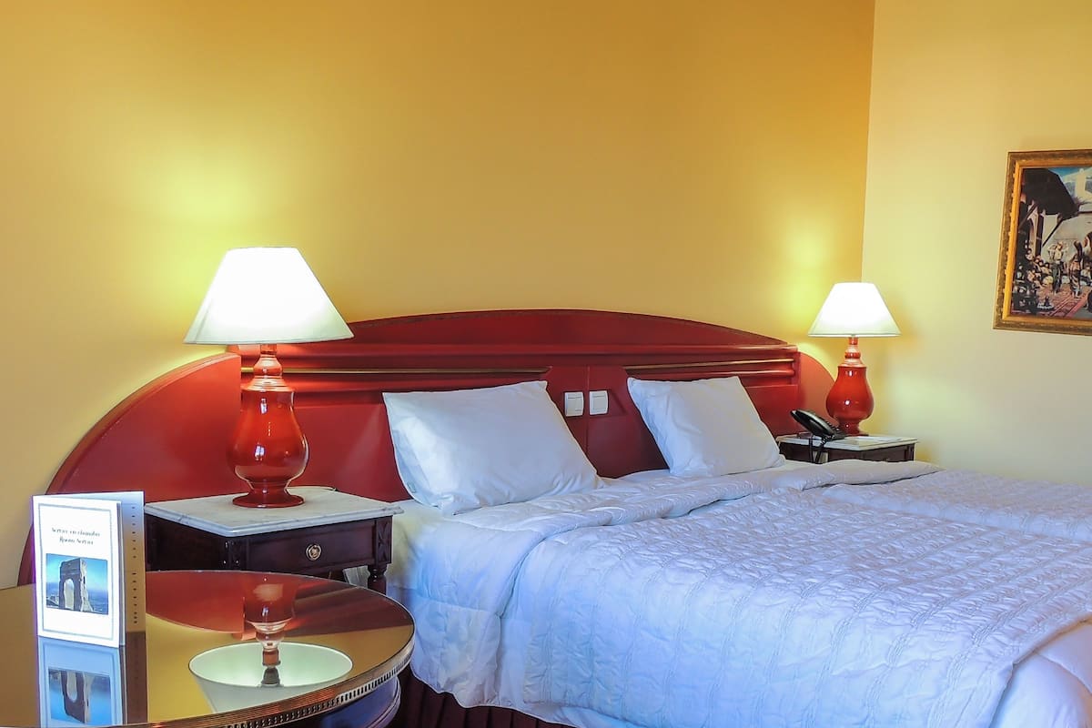 A hotel room with a red headboard, two bedside lamps, a bed with white bedding, a nightstand with a lamp, a framed picture on the wall, and a brochure on the side table.