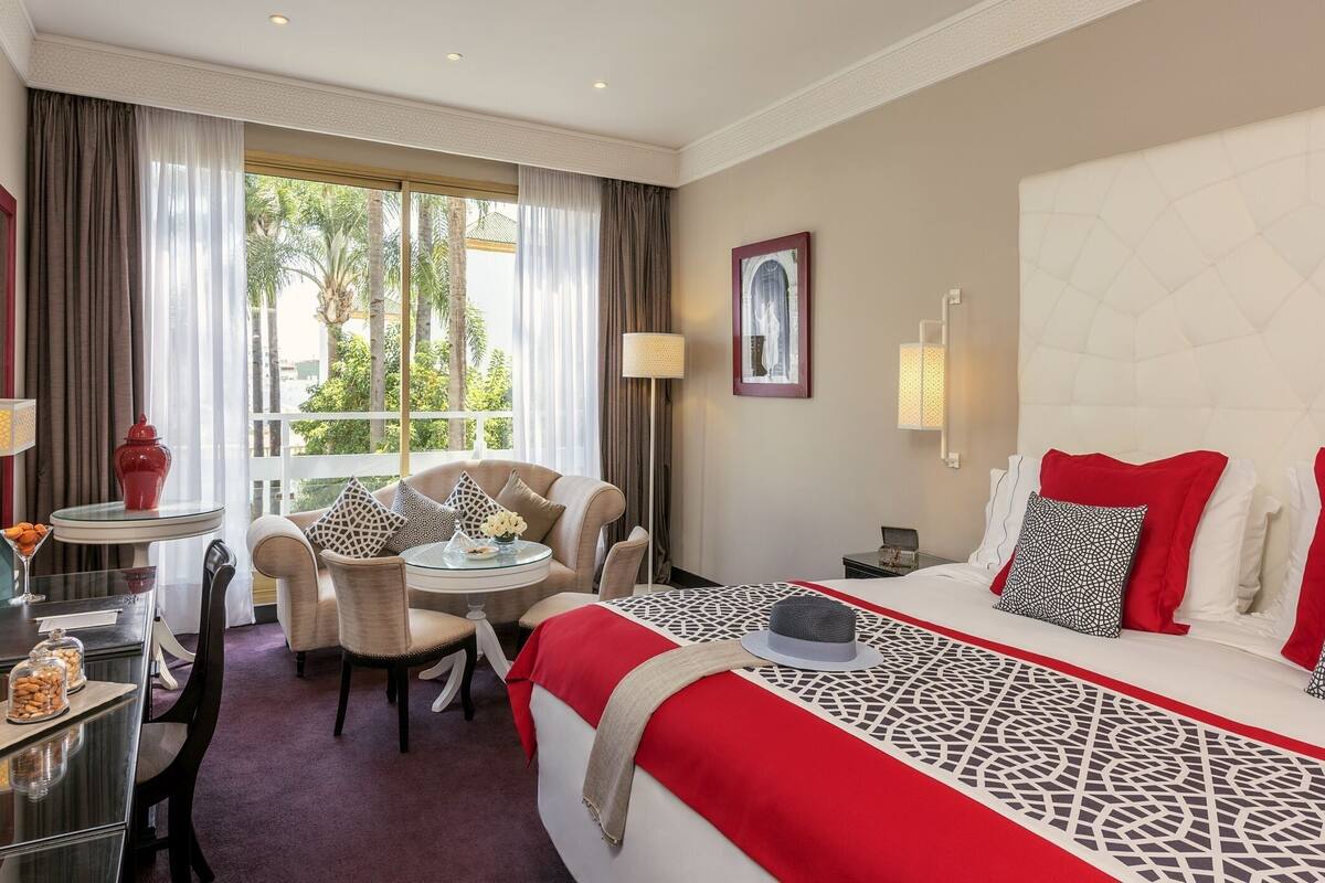 A hotel room with a bed, a seating area with a table and chairs, a desk with a lamp, and a view of palm trees through the window.
