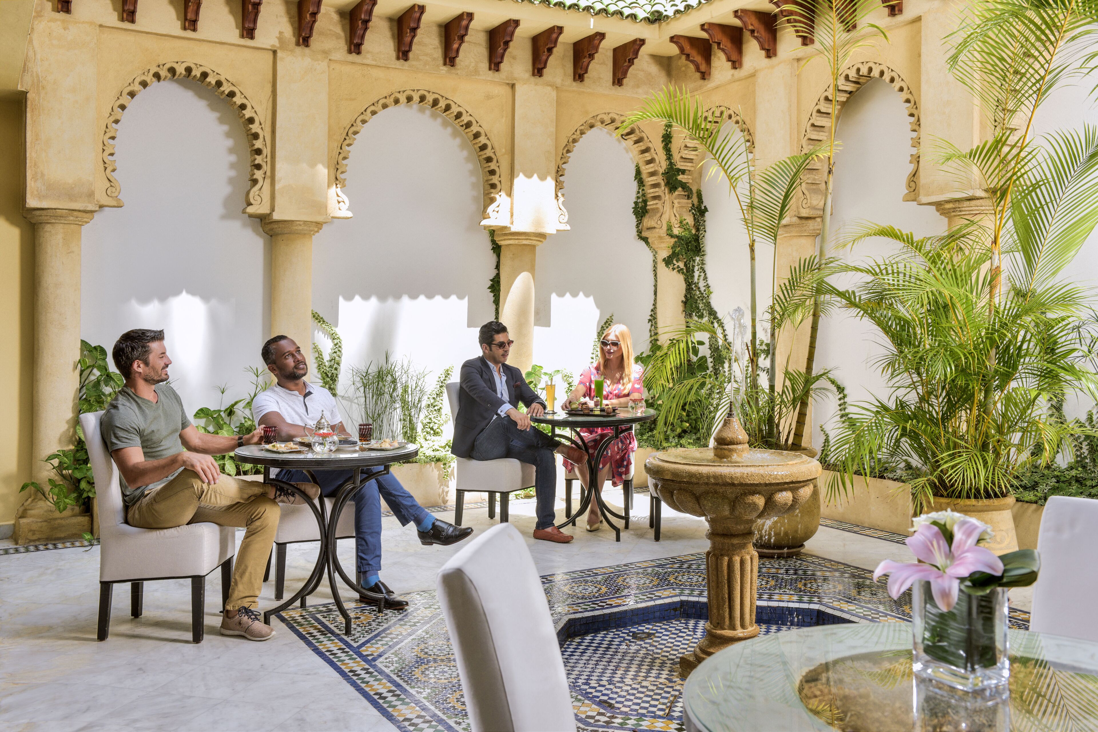 A group of people seated at a round table in a courtyard with arches and potted plants.