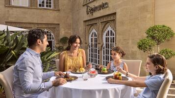 Un grupo de personas cenando en una mesa al aire libre con mantel blanco, rodeadas por un edificio con ventanas ornamentadas y un letrero que dice "Taj Mahal".