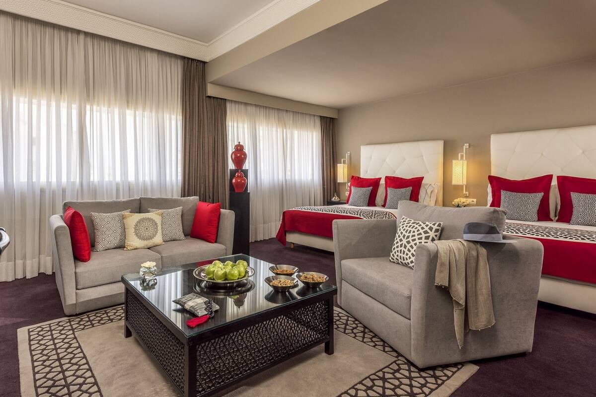 A well-appointed hotel room with a sofa, armchair, and a bed with red bedding. A coffee table with fruits and a vase of flowers is in the center.