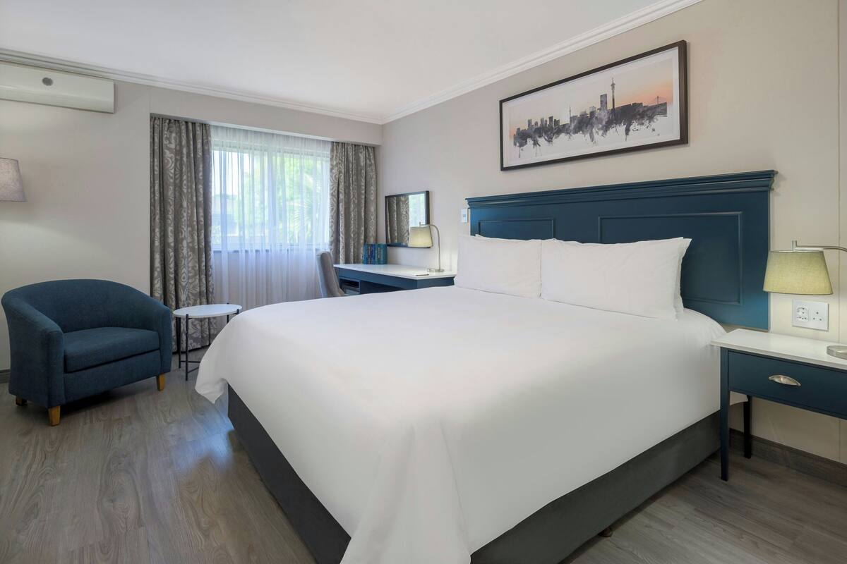 A hotel room with a large bed, a desk, a chair, and a cityscape artwork on the wall.