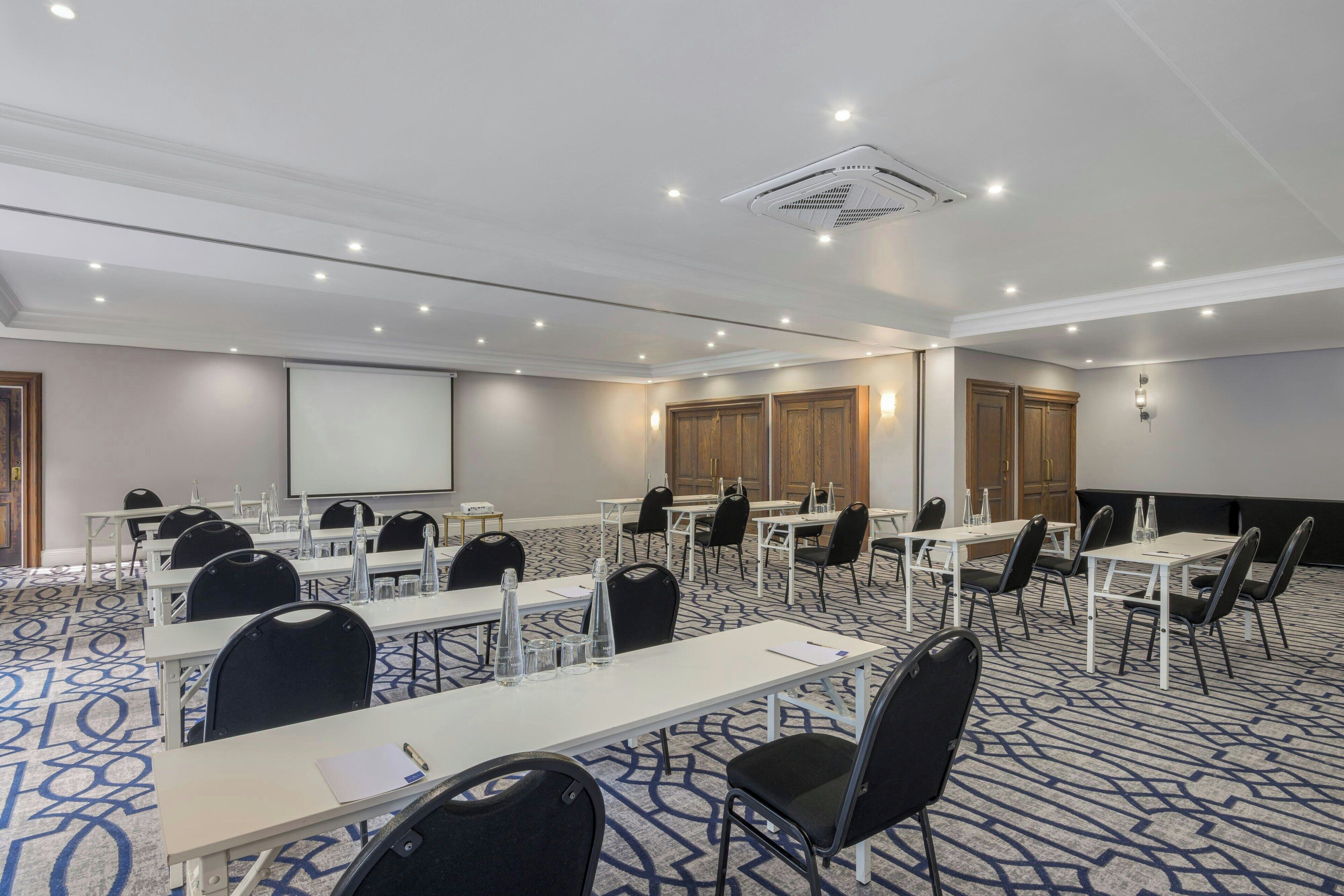 Un salón de conferencias con mesas, sillas, una pantalla de proyector y suelo alfombrado.