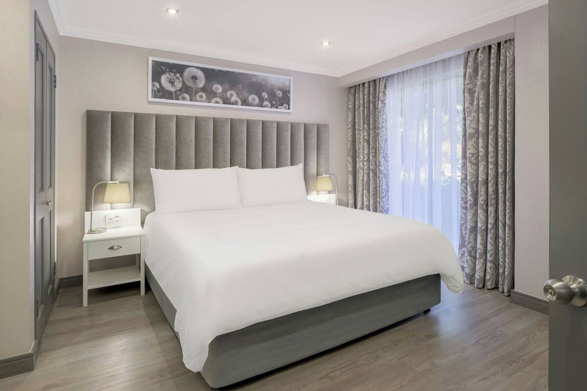 A modern hotel room with a large bed, two bedside tables with lamps, a headboard, and a large window with curtains.