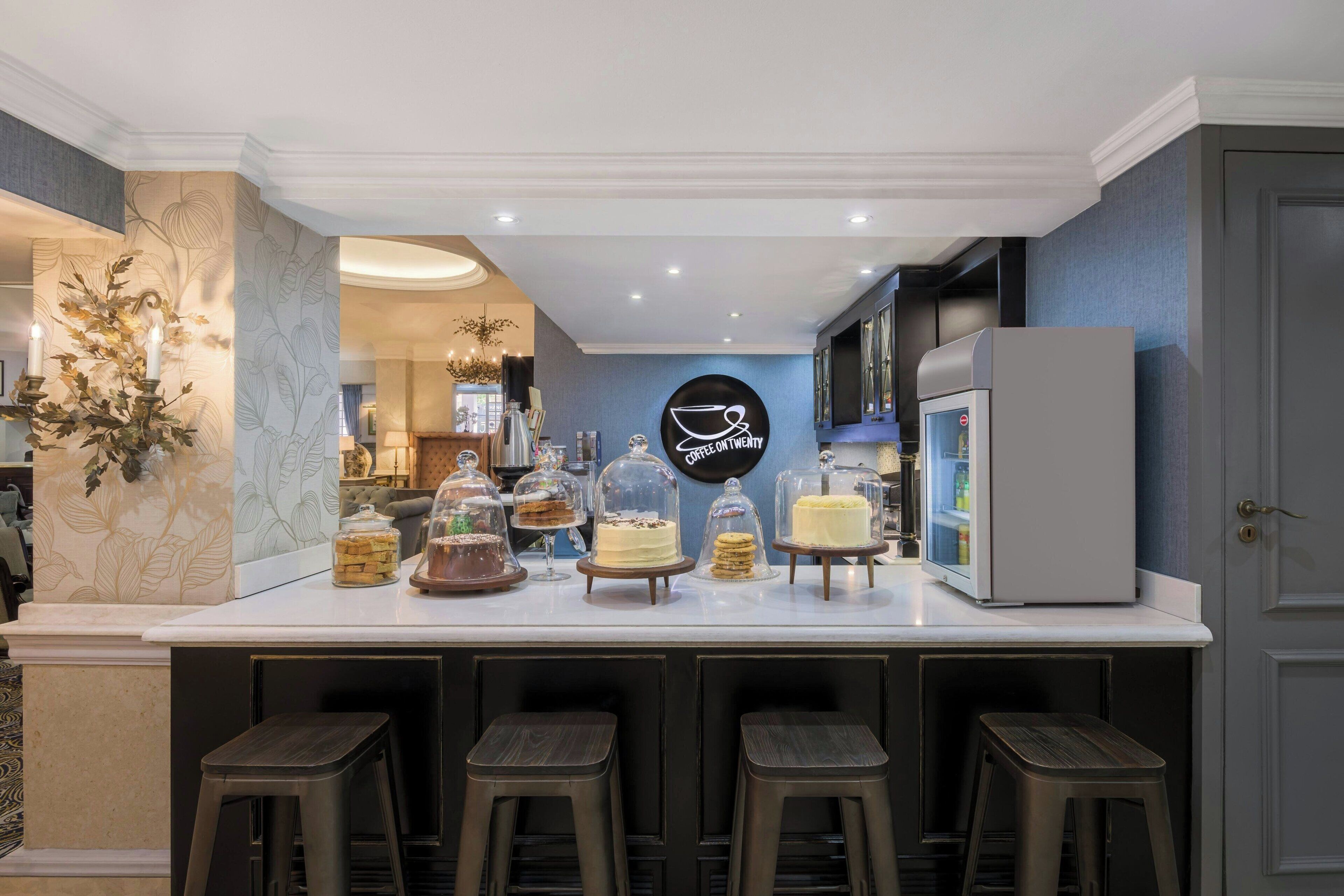 Un espace bar moderne avec un comptoir blanc, de nombreux tabourets et une variété de desserts présentés sous des cloches de verre.