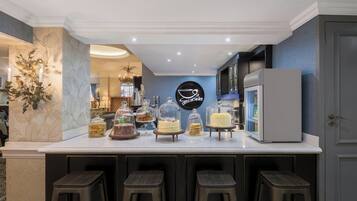 Un espace bar moderne avec un comptoir blanc, de nombreux tabourets et une variété de desserts présentés sous des cloches de verre.