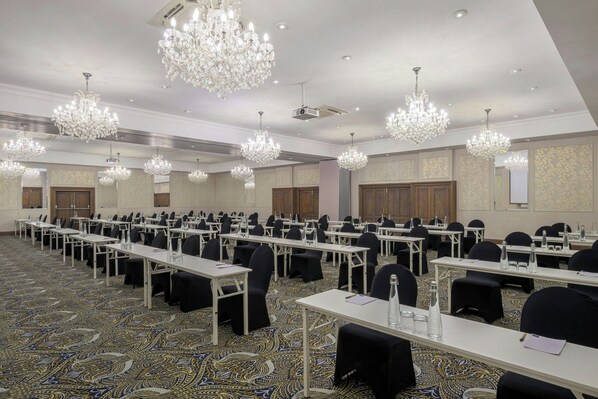 A conference room with rows of tables, black chairs, and ornate chandeliers.