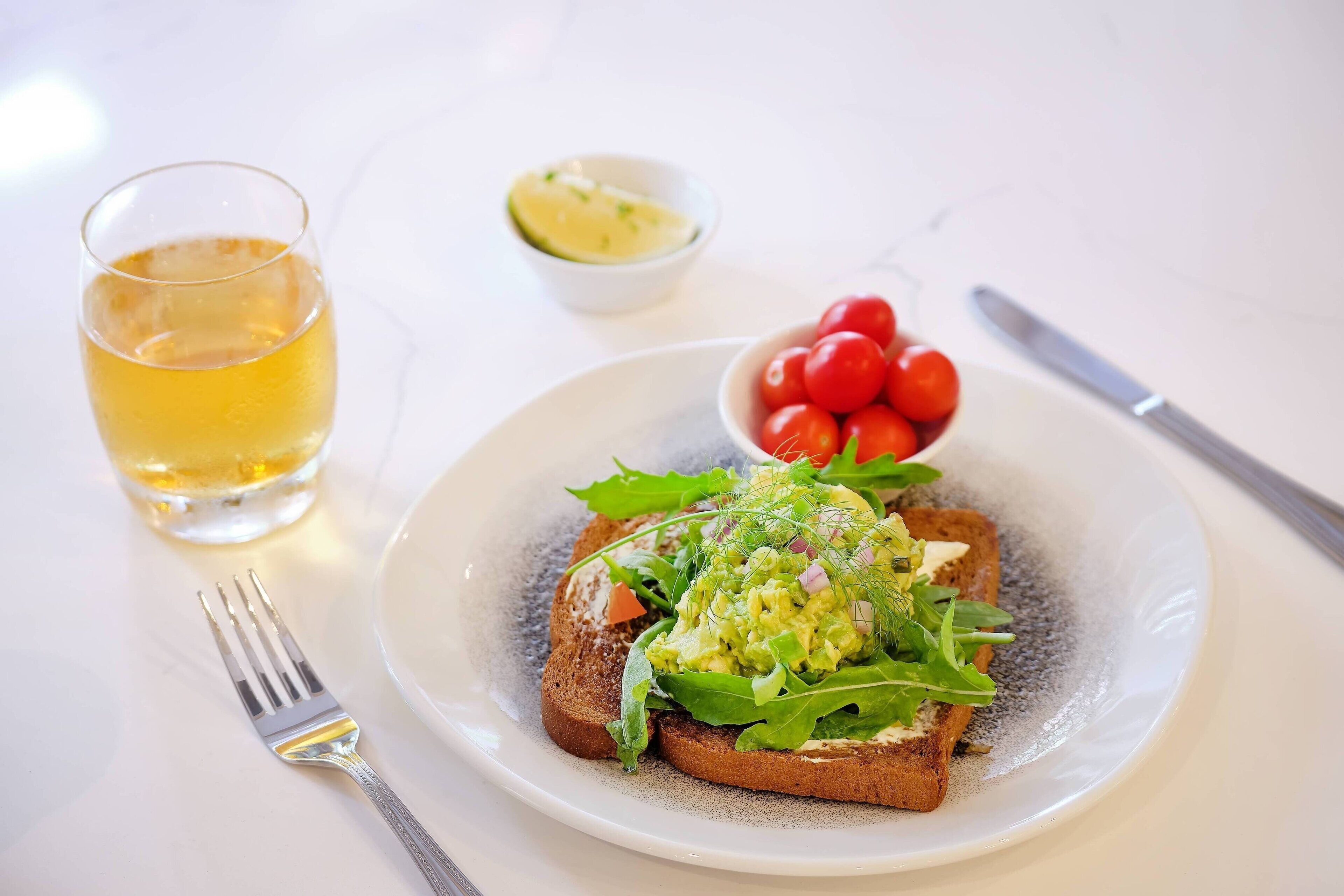 Un plato con tostadas cubiertas de aguacate y verduras, un bol pequeño de tomates cherry y un vaso de bebida amarilla sobre una mesa blanca.