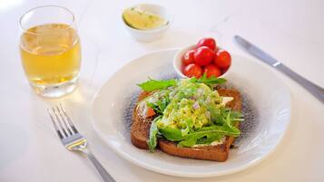 Un plato con tostadas cubiertas de aguacate y verduras, un bol pequeño de tomates cherry y un vaso de bebida amarilla sobre una mesa blanca.