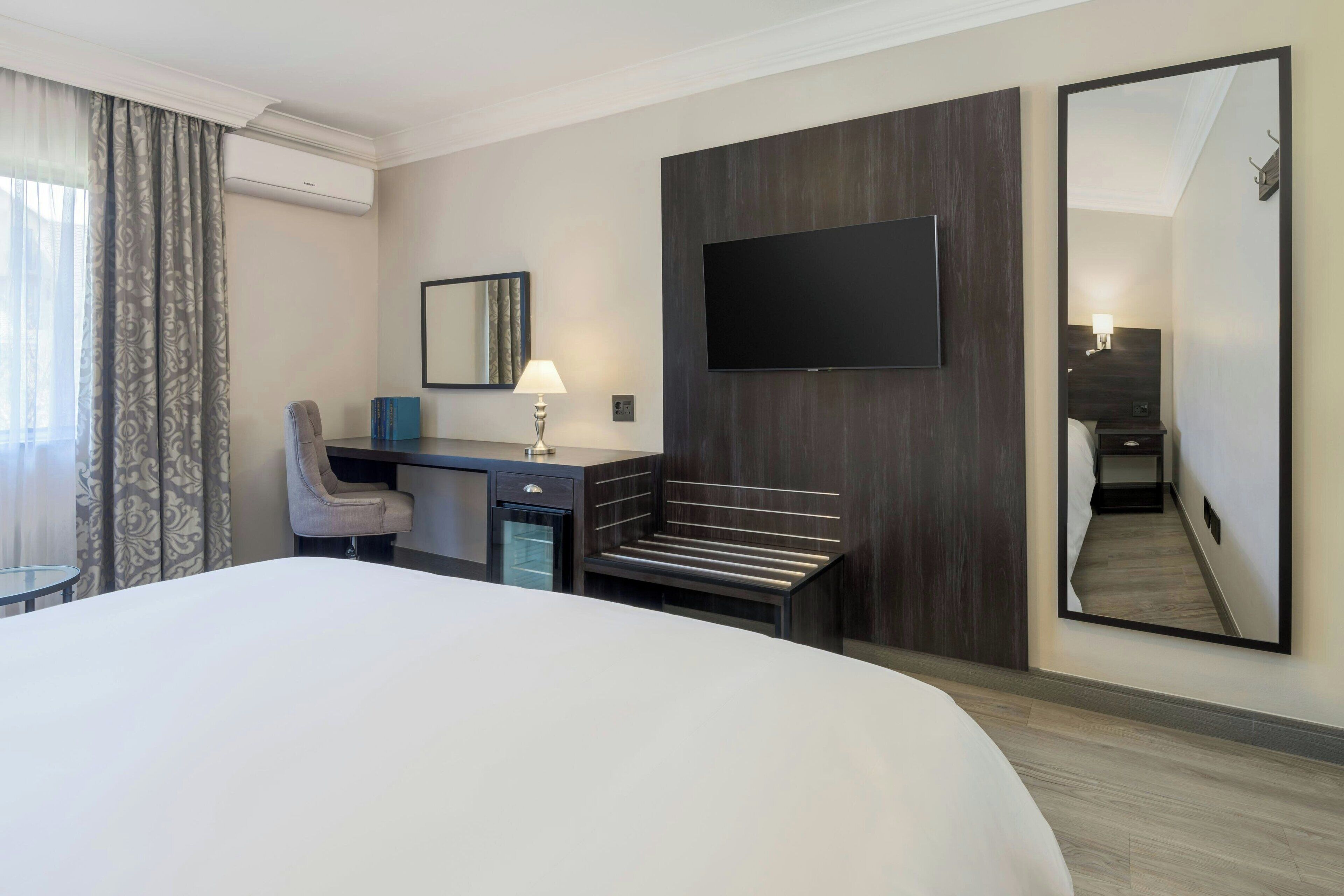 A modern hotel room with a large bed, a desk with a chair, a TV mounted on the wall, and a large mirror.