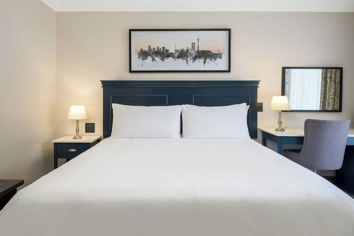 A hotel room with a large bed, two bedside tables with lamps, a chair, and a cityscape artwork on the wall.