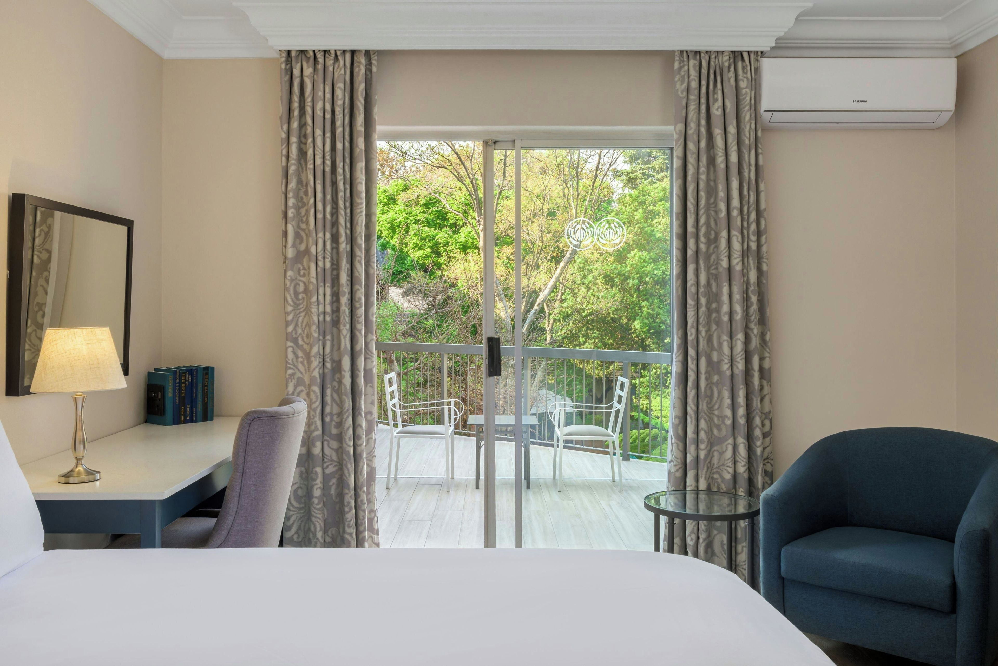 Quarto de hotel com cama, escrivaninha com abajur, cadeira, poltrona azul, varanda com mesa e cadeiras, e vista para o verde.
