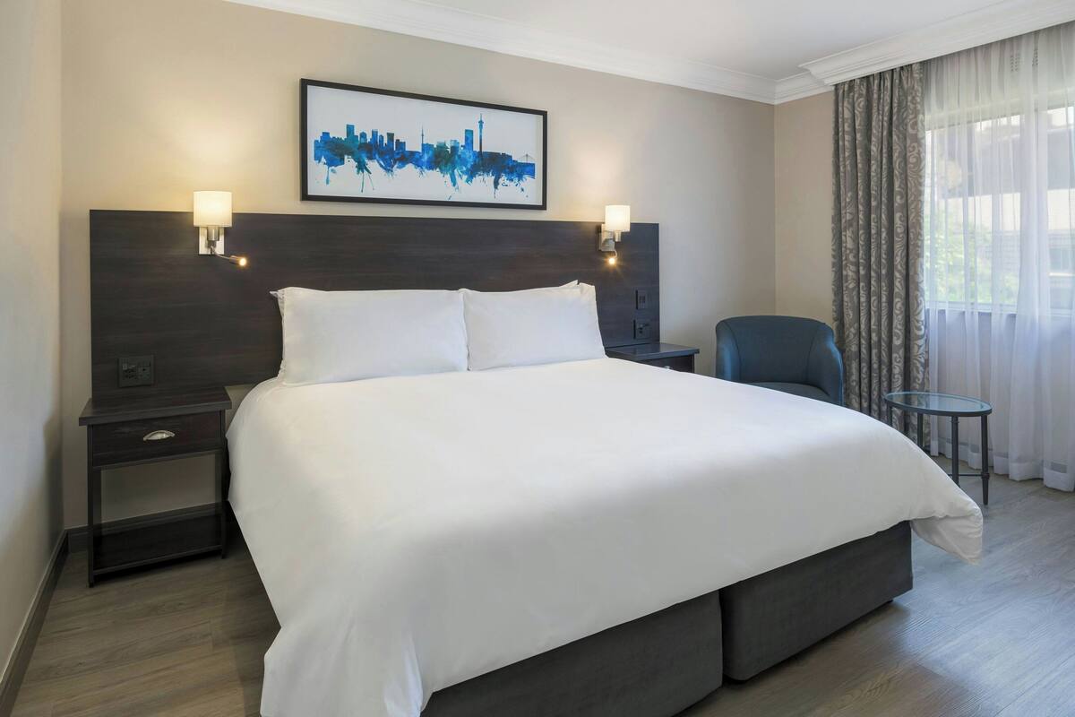 A hotel room with a large bed, two bedside tables, a chair, a small table, and a cityscape artwork on the wall.