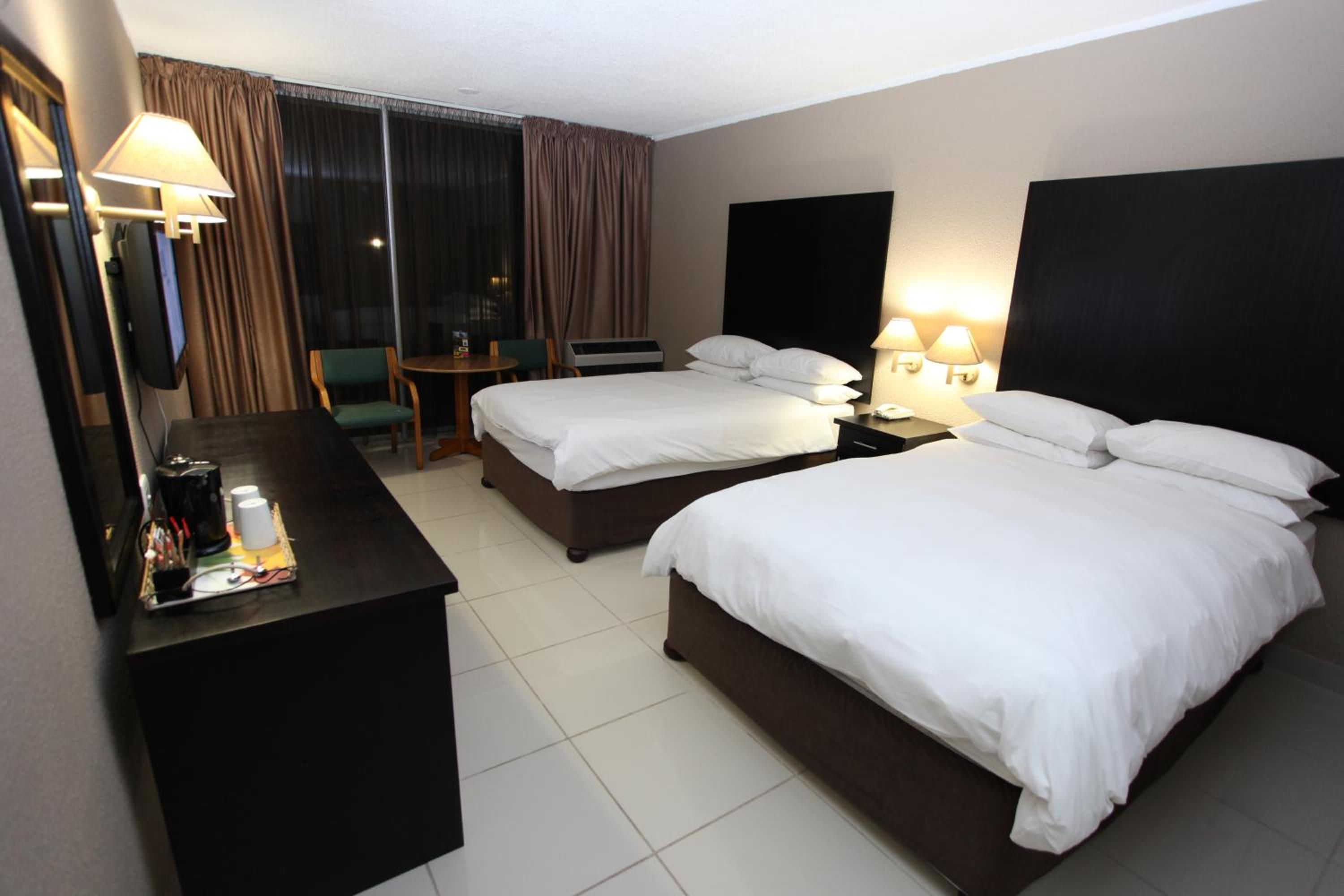 Ein Hotelzimmer mit zwei Betten, einem Schreibtisch mit Lampe, einem Sessel, einem Fernseher und einem fenster mit Vorhängen.