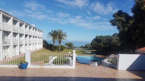 Un área de piscina con vista al océano, un edificio blanco con balcones y una palmera.