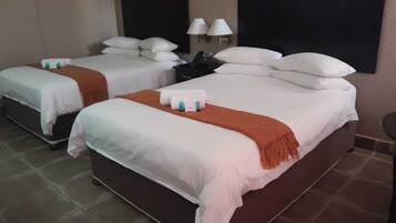 Une chambre d’hôtel avec deux lits, chacun recouvert de draps blancs et d’un plaid marron. Il y a deux tables de chevet avec des lampes et une petite table avec un téléphone.