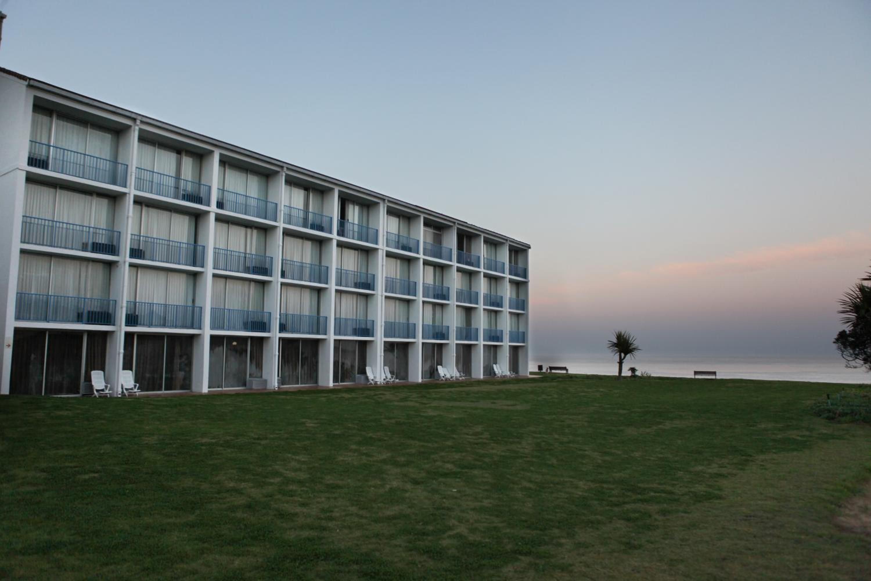 Un hotel de varios pisos con balcones, un césped y vista al mar.