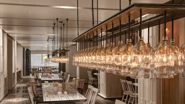 A spacious dining area with long tables, hanging light fixtures, and a view of the kitchen in the background. - Gloria Prince Hotel Taipei (Taipei)