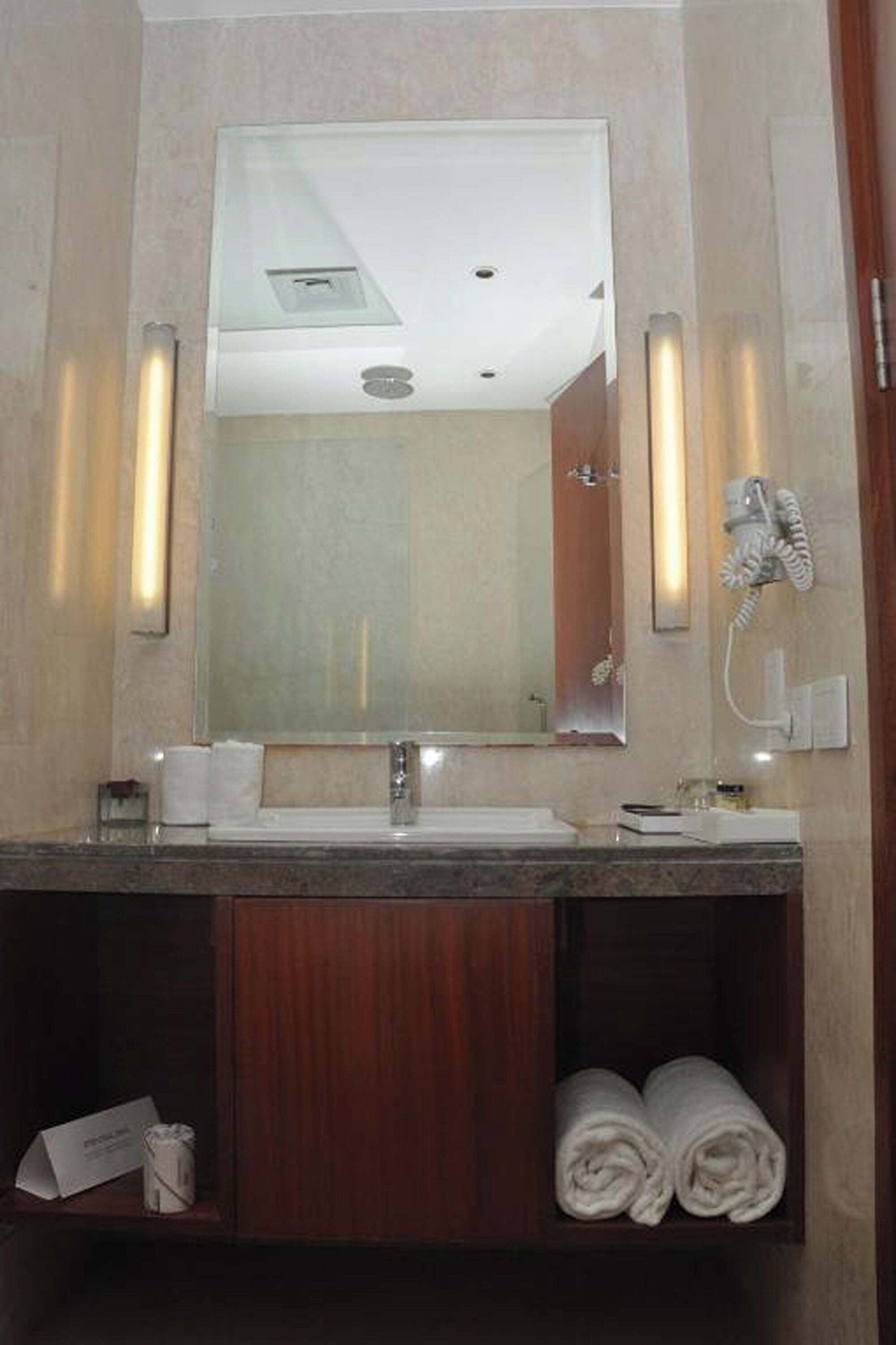 Un baño de hotel con un espejo grande, dos lavamanos, un perchero para toallas y un secador de pelo.
