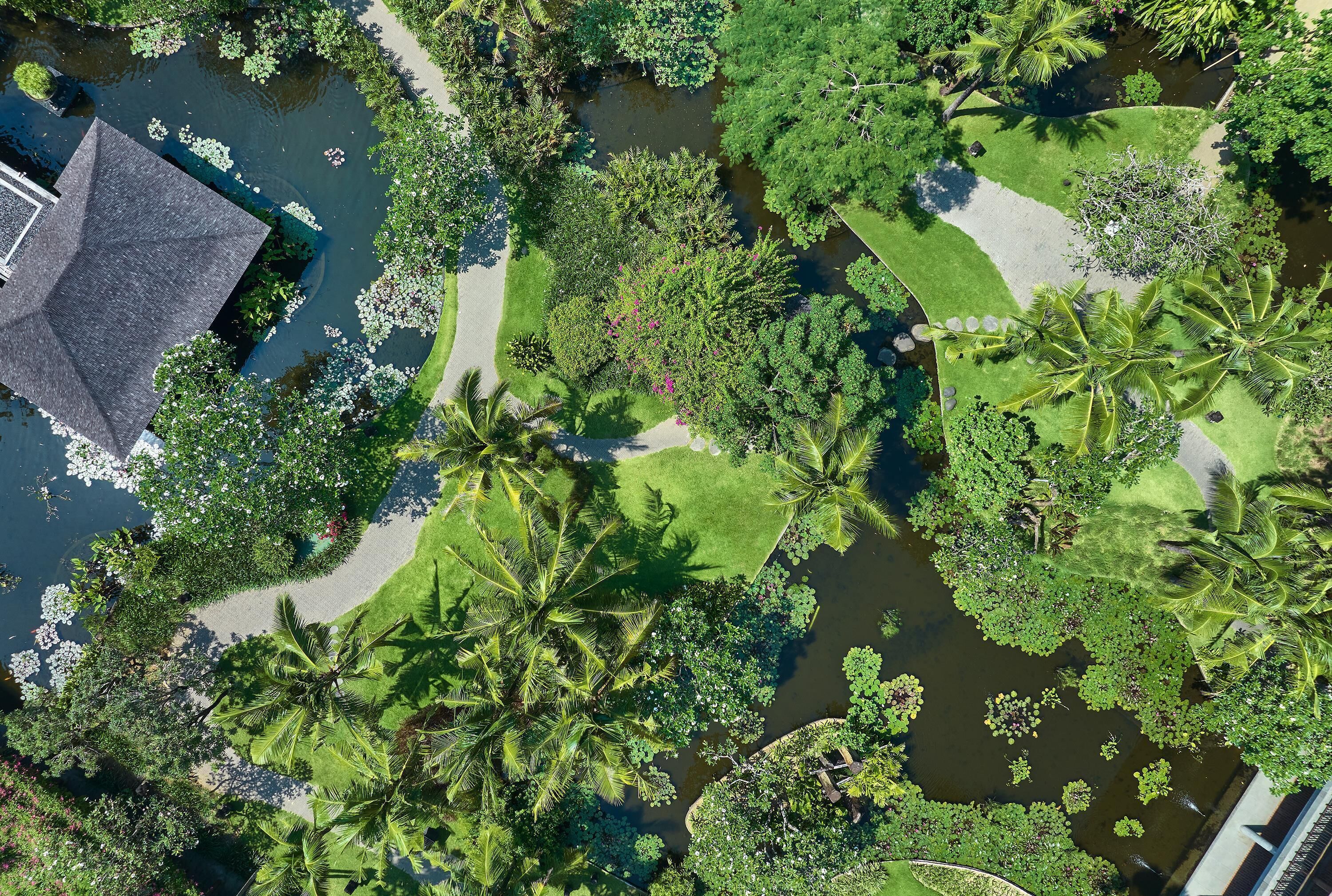 An aerial view of a landscaped area with a pond, palm trees, and a building with a dark roof.