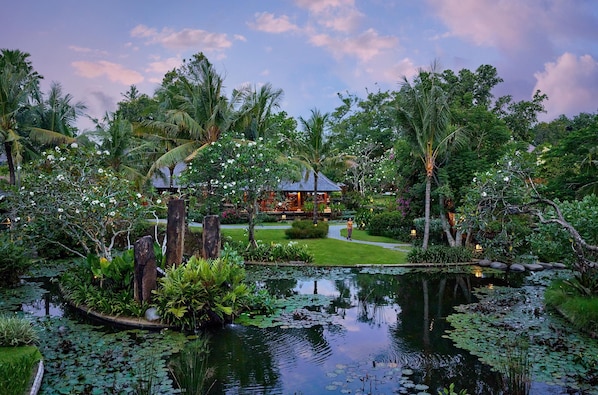 A tropical garden with a pond, lush greenery, and a small building with a thatched roof.