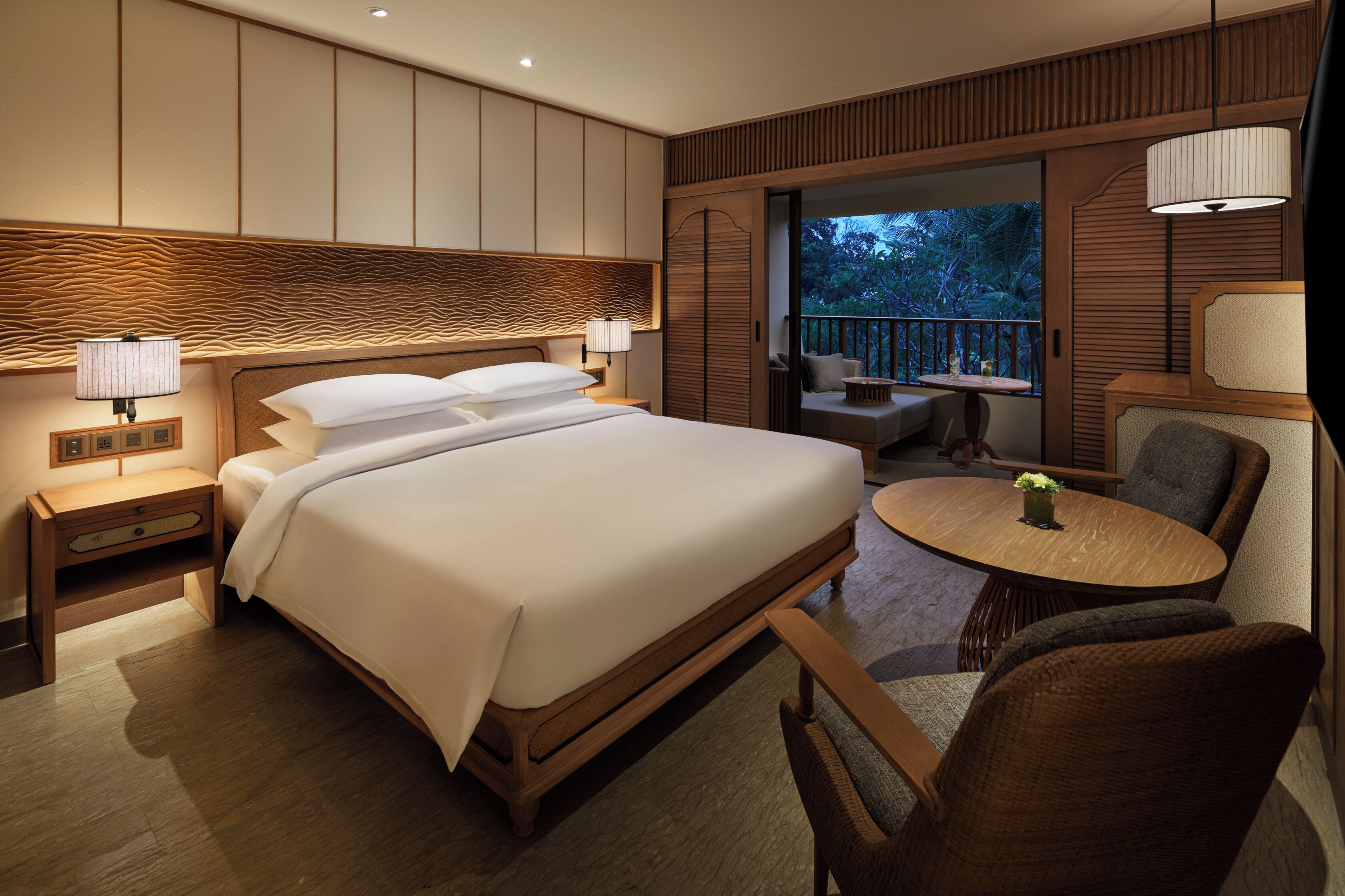 A modern hotel room with a large bed, a round dining table, and a balcony with a view of the outdoors.