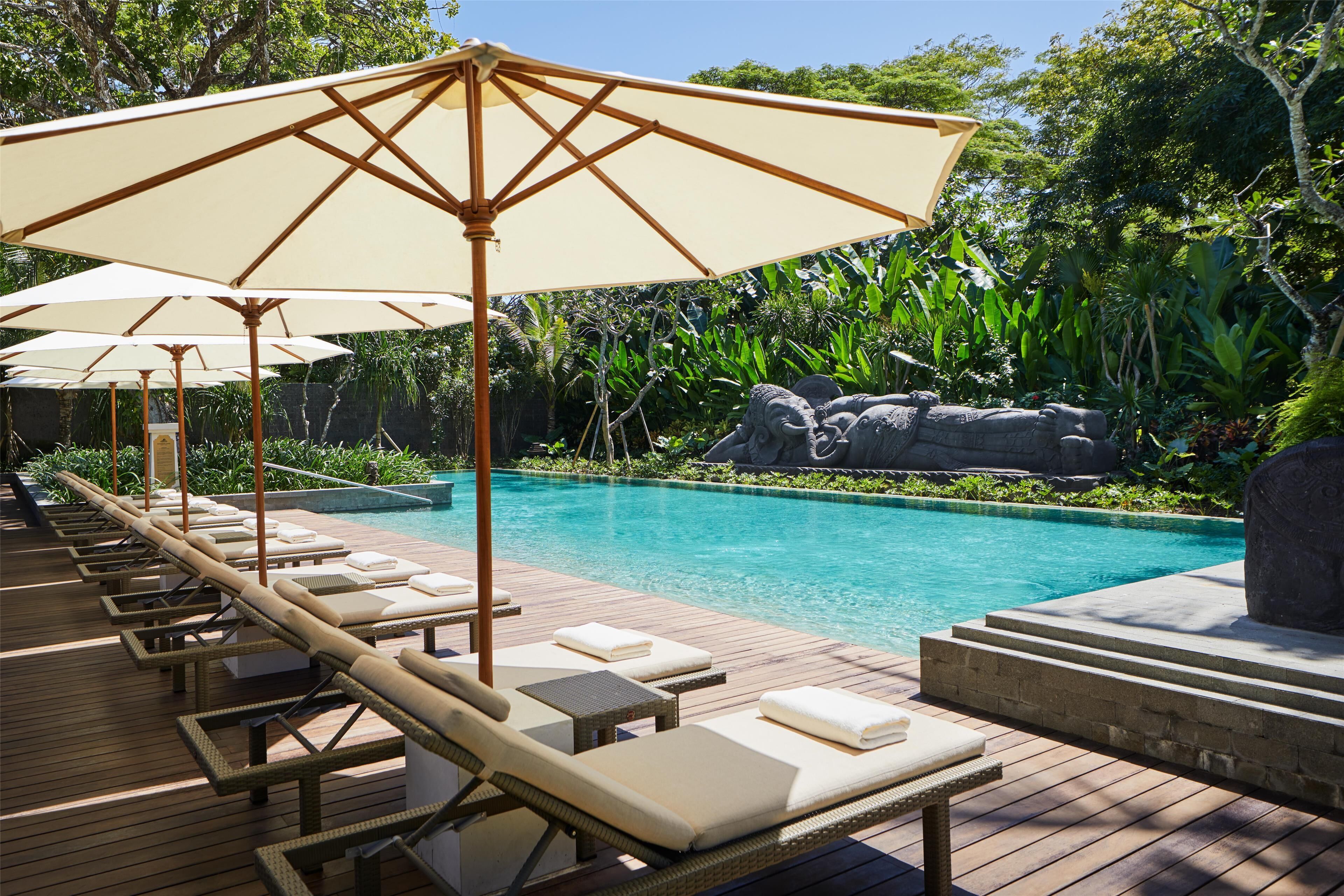 A poolside area with lounge chairs and umbrellas, surrounded by lush greenery and a rocky backdrop.